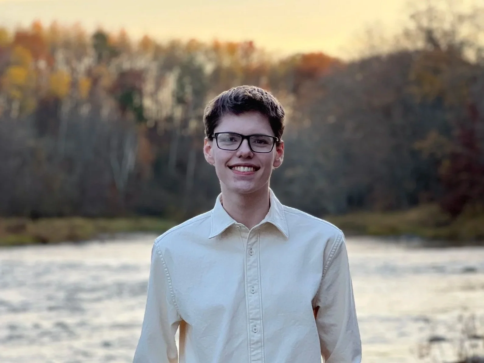 A young man standing outdoors in front of a river with trees in autumn colors, smiling at the camera.