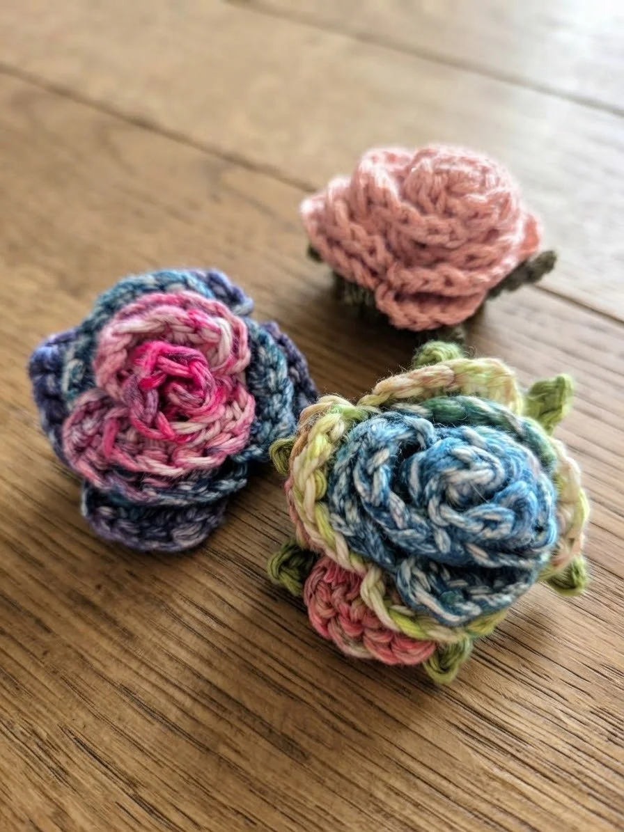 Three small crochet roses in pink, blue, and multicolored yarns on a wooden surface.