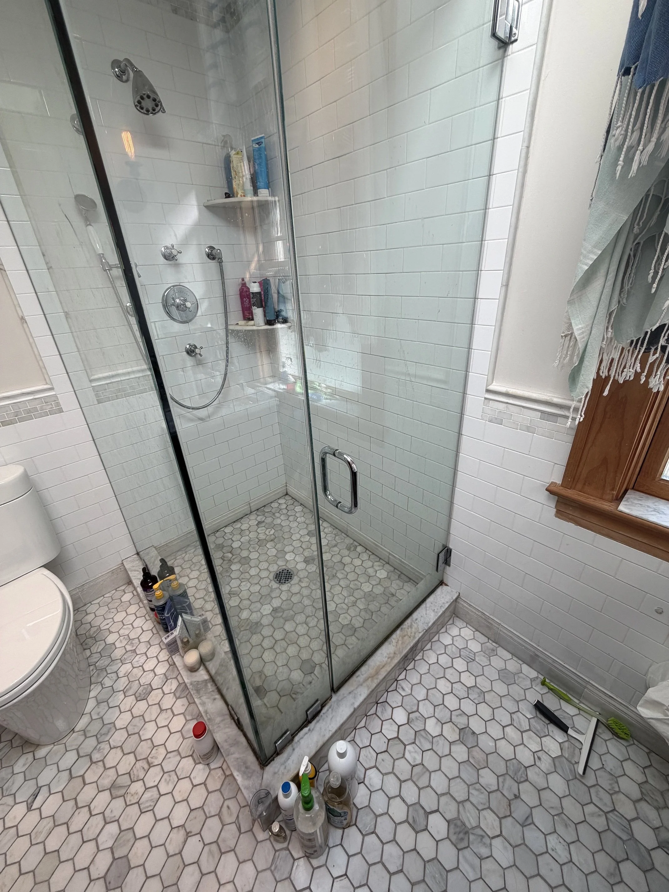 Glass-enclosed bathroom shower with white subway tile walls, hexagonal floor tiles, chrome fixtures, and toiletries