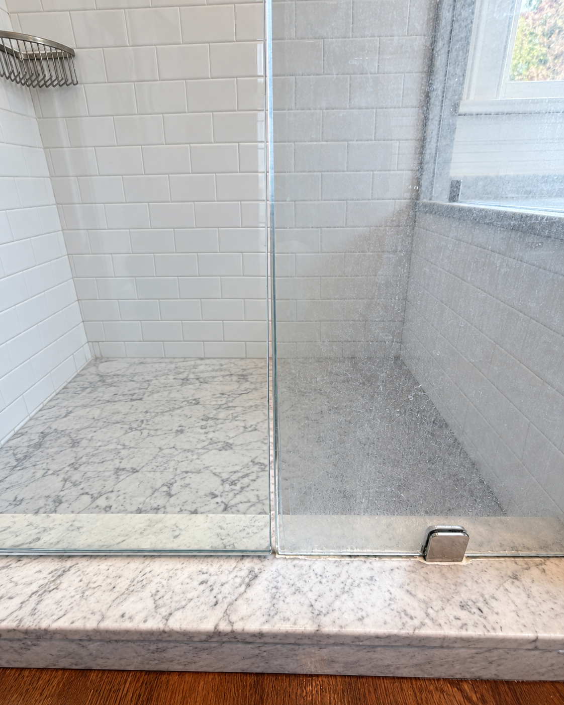 Glass-enclosed shower with white subway tile walls, marble threshold, rain-speckled glass door, and metal corner shelf