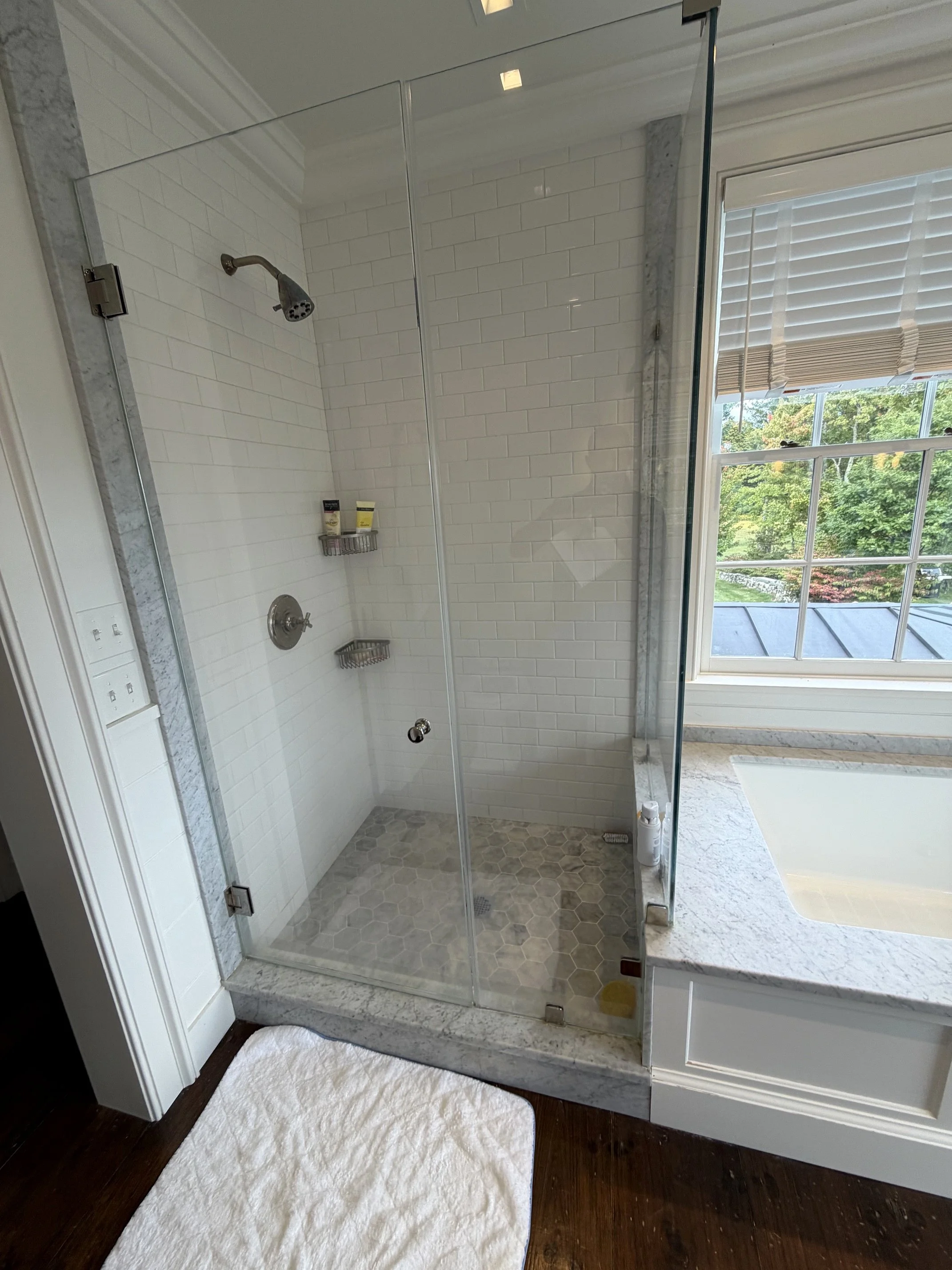 Modern bathroom with white subway tile walk-in shower, glass doors, marble trim, and adjacent bathtub beneath a window.