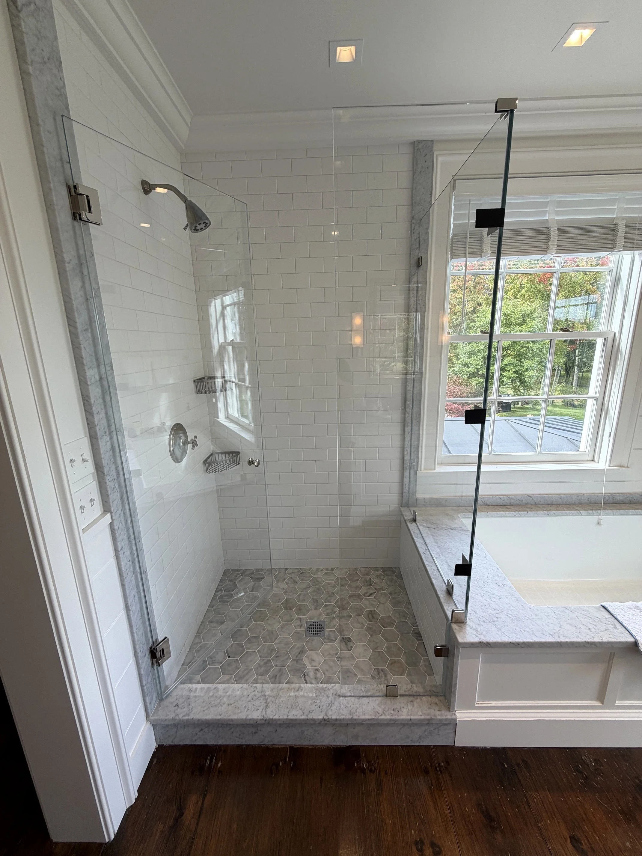 Walk-in bathroom shower with white subway tile walls, marble trim, hexagon floor tiles, glass doors, and a rainfall showerhead beside a bathtub window.
