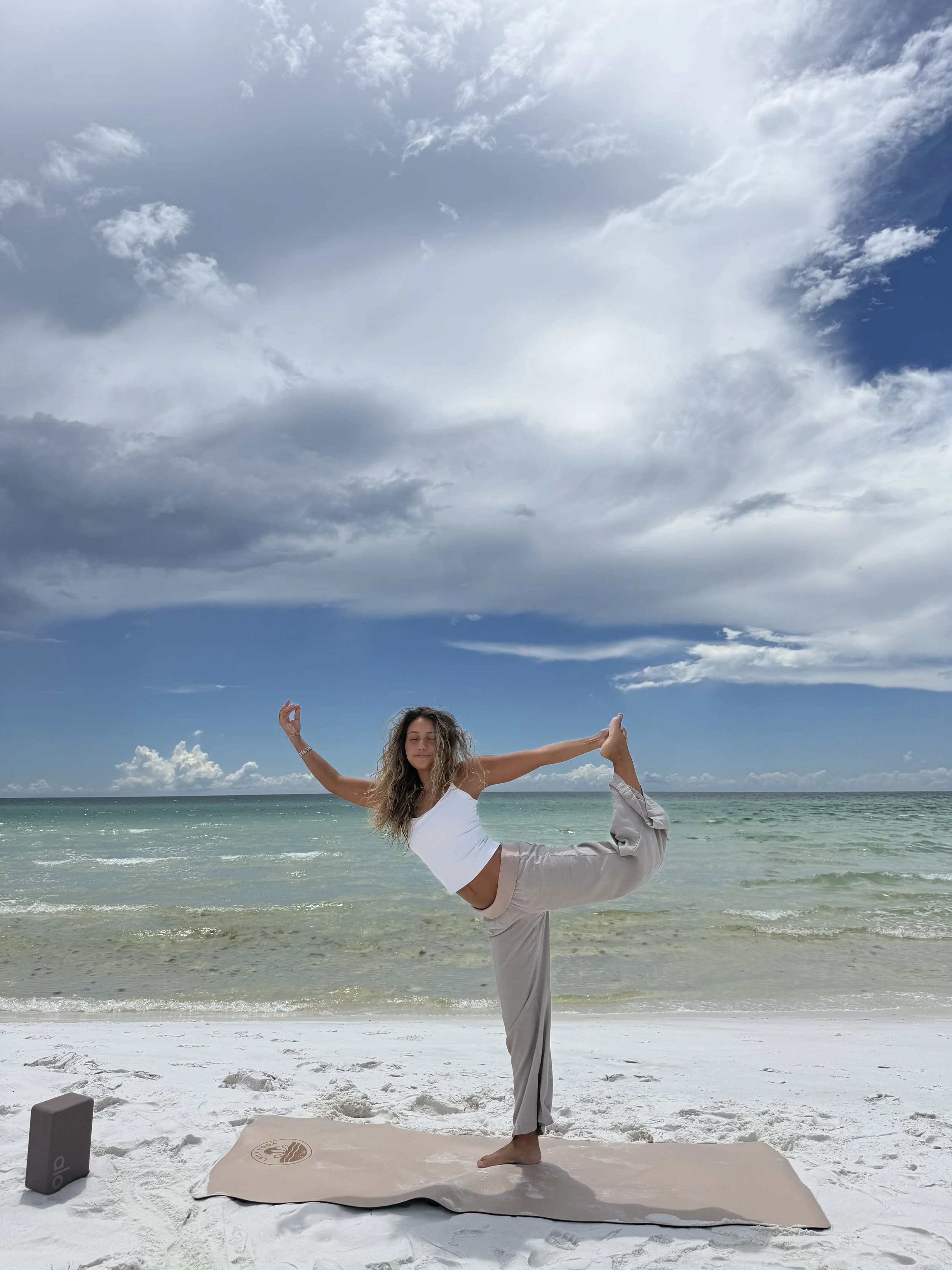 A woman practicing yoga on a beach, standing on one leg in a yoga pose, with a yoga mat, a block, and a rolled-up towel nearby, ocean in the background and cloudy sky overhead.