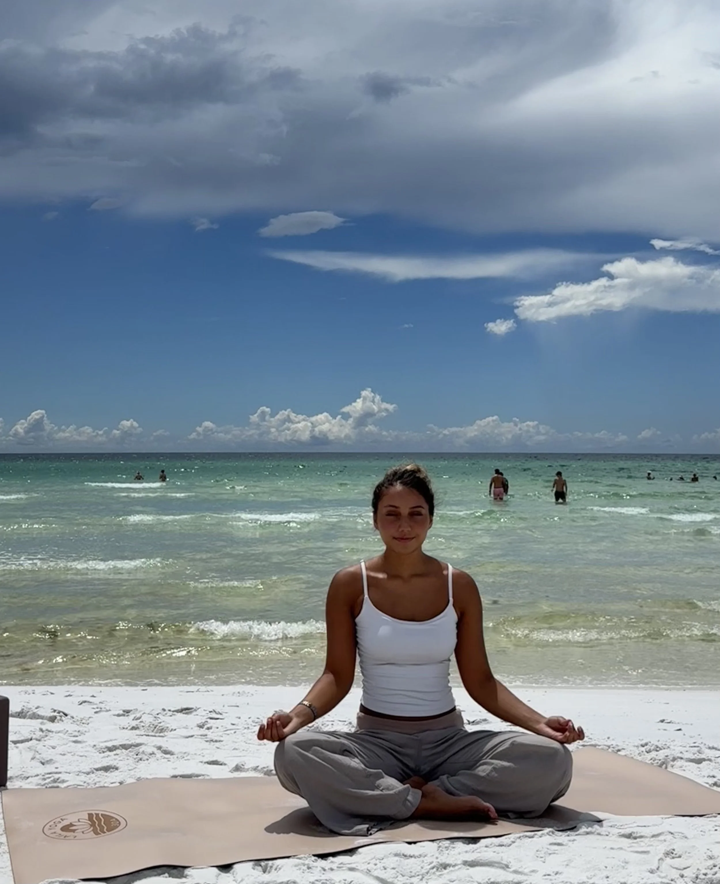 A woman practicing meditation on a beach, sitting cross-legged on a yoga mat with her hands in a mudra position, facing the ocean with a cloudy sky above and other people swimming in the water.