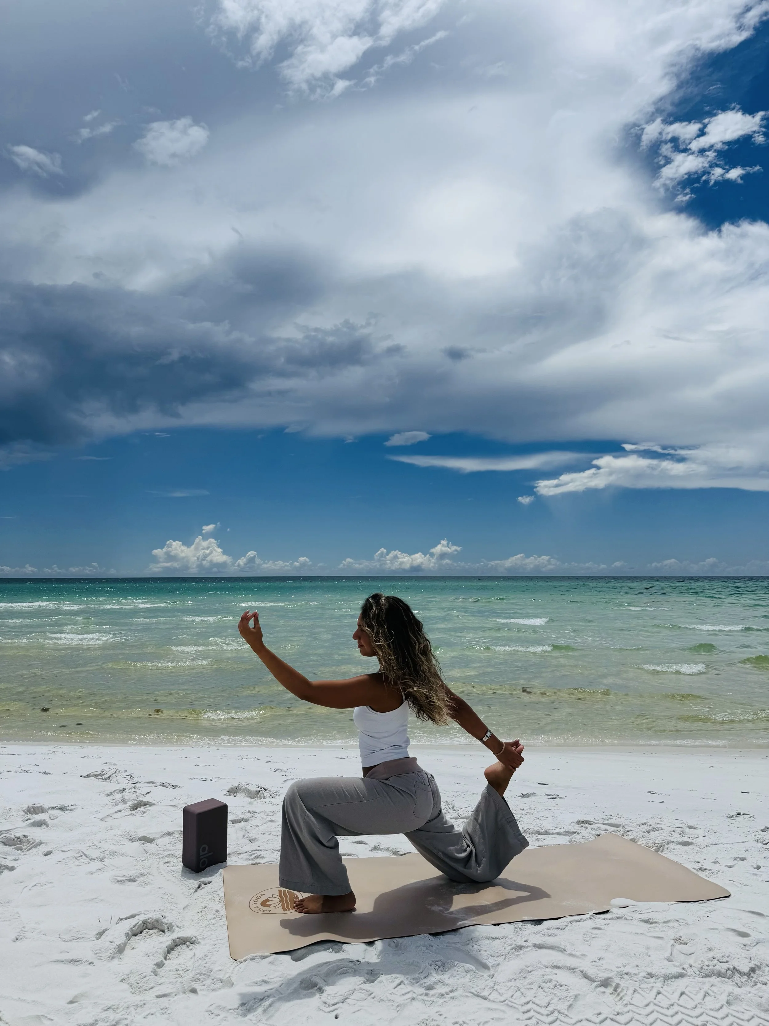 A woman practicing yoga on a beach with ocean waves, clouds in the sky, and a yoga mat. She is in a lunge pose with one arm reaching upward and the other holding her back foot.