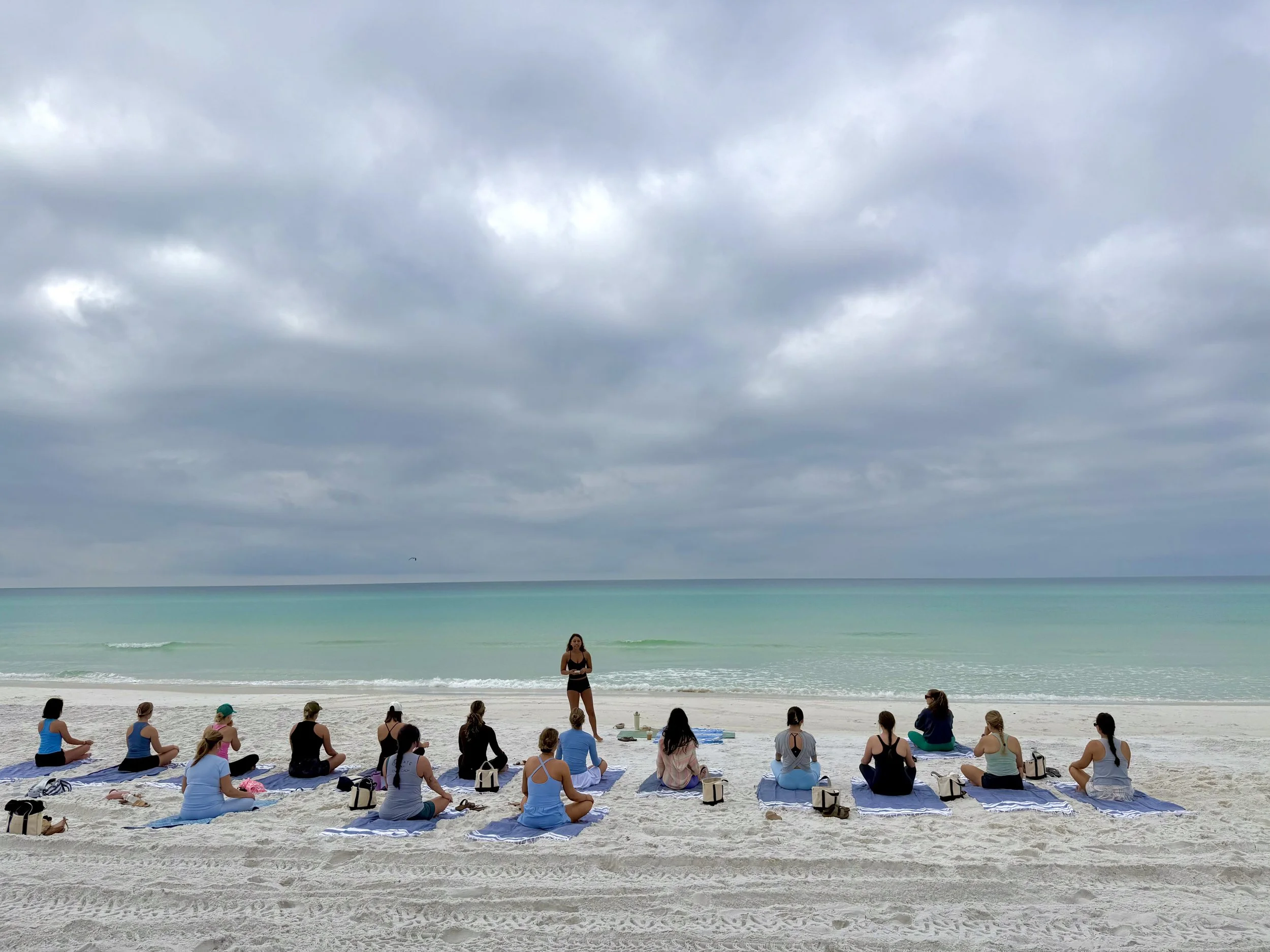 A group of people participating in a yoga or meditation class on a sandy beach. An instructor stands in front of the class, which is facing the ocean under a cloudy sky. The participants are seated on towels, focusing on the instructor, with the ocean and cloudy sky in the background.