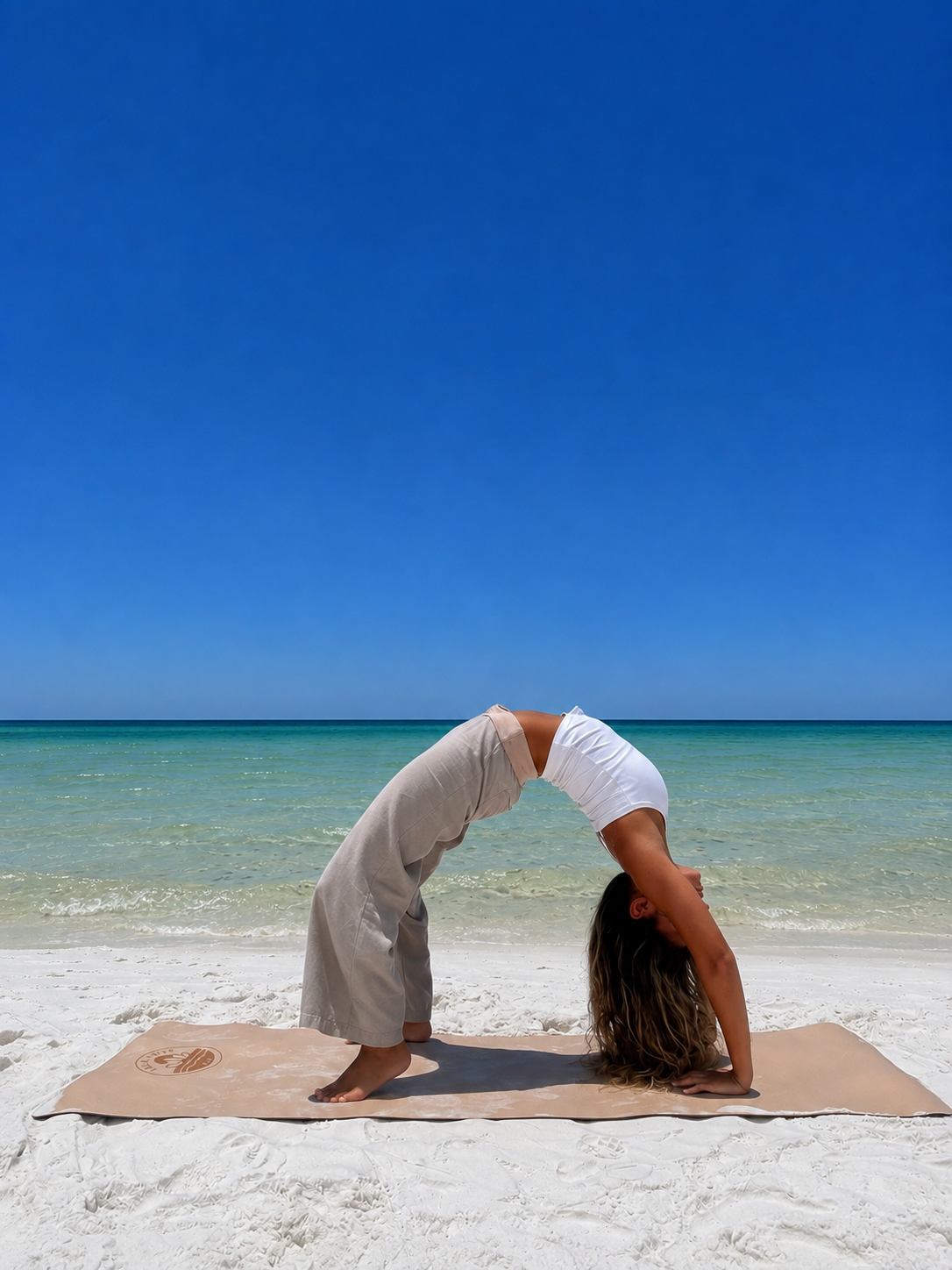 A woman practicing yoga on a beach, performing a backbend pose on a mat with ocean and clear sky in the background.