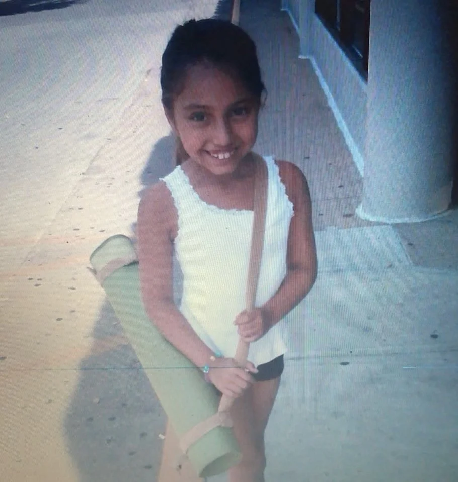 A young girl smiling, wearing a white sleeveless top, carrying a rolled-up yoga mat or foam roller over her shoulder with a strap, standing on a sidewalk outside a building.