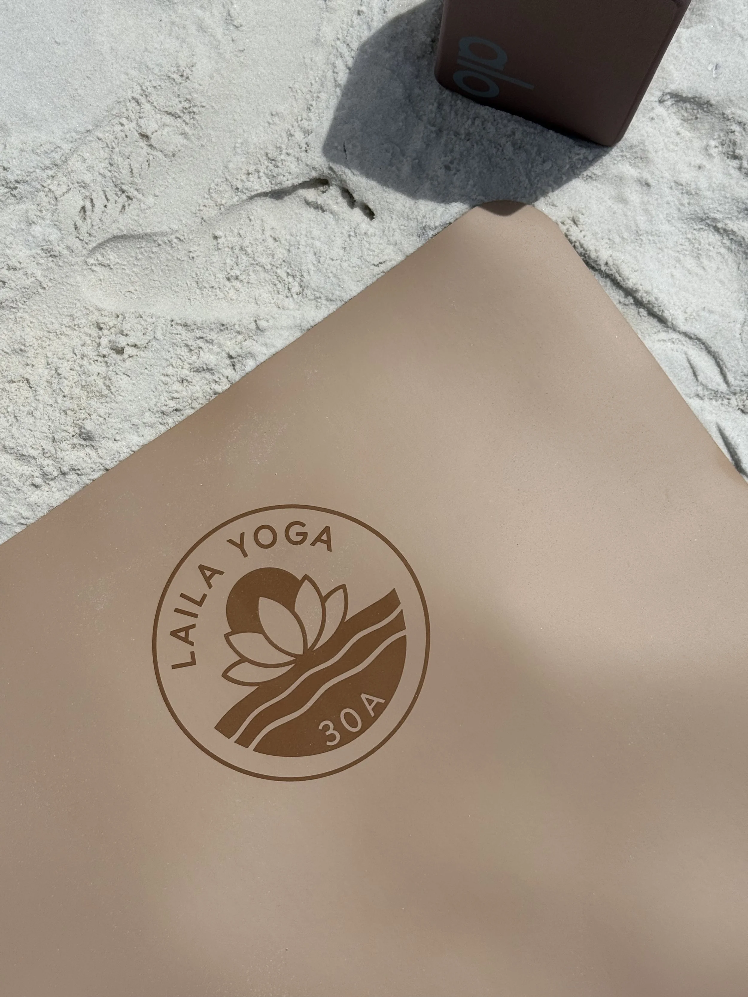 A yoga mat with a logo that reads 'LA YOGA' in a circle, placed on a sandy surface next to a cardboard box.