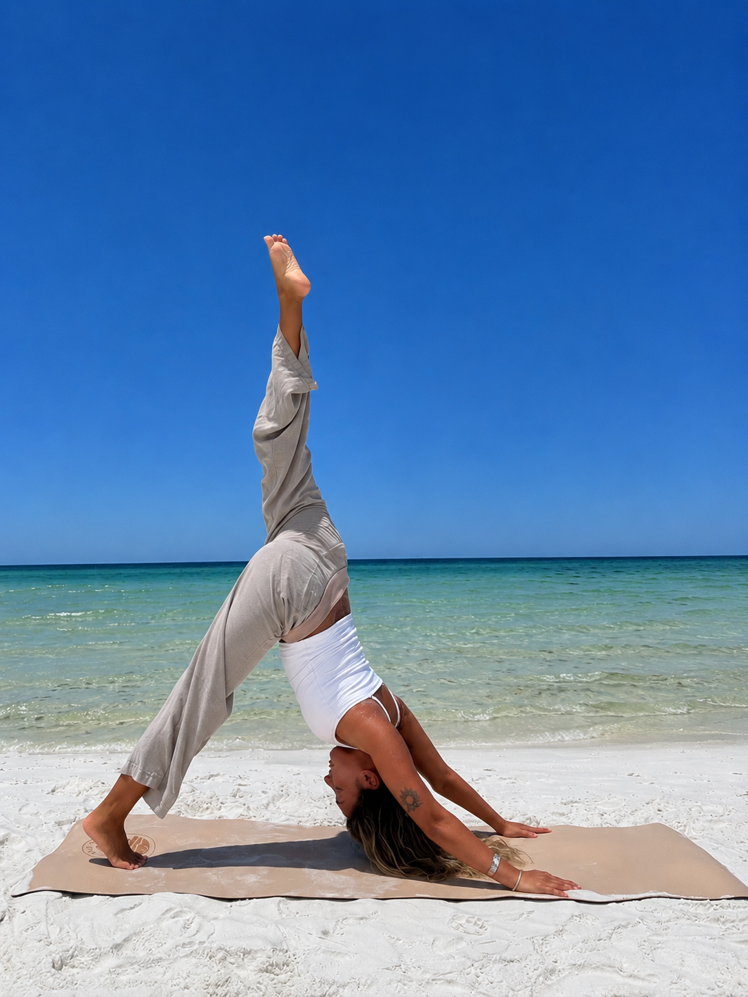 Woman practicing yoga in downward dog pose on the beach with the ocean and blue sky in the background.