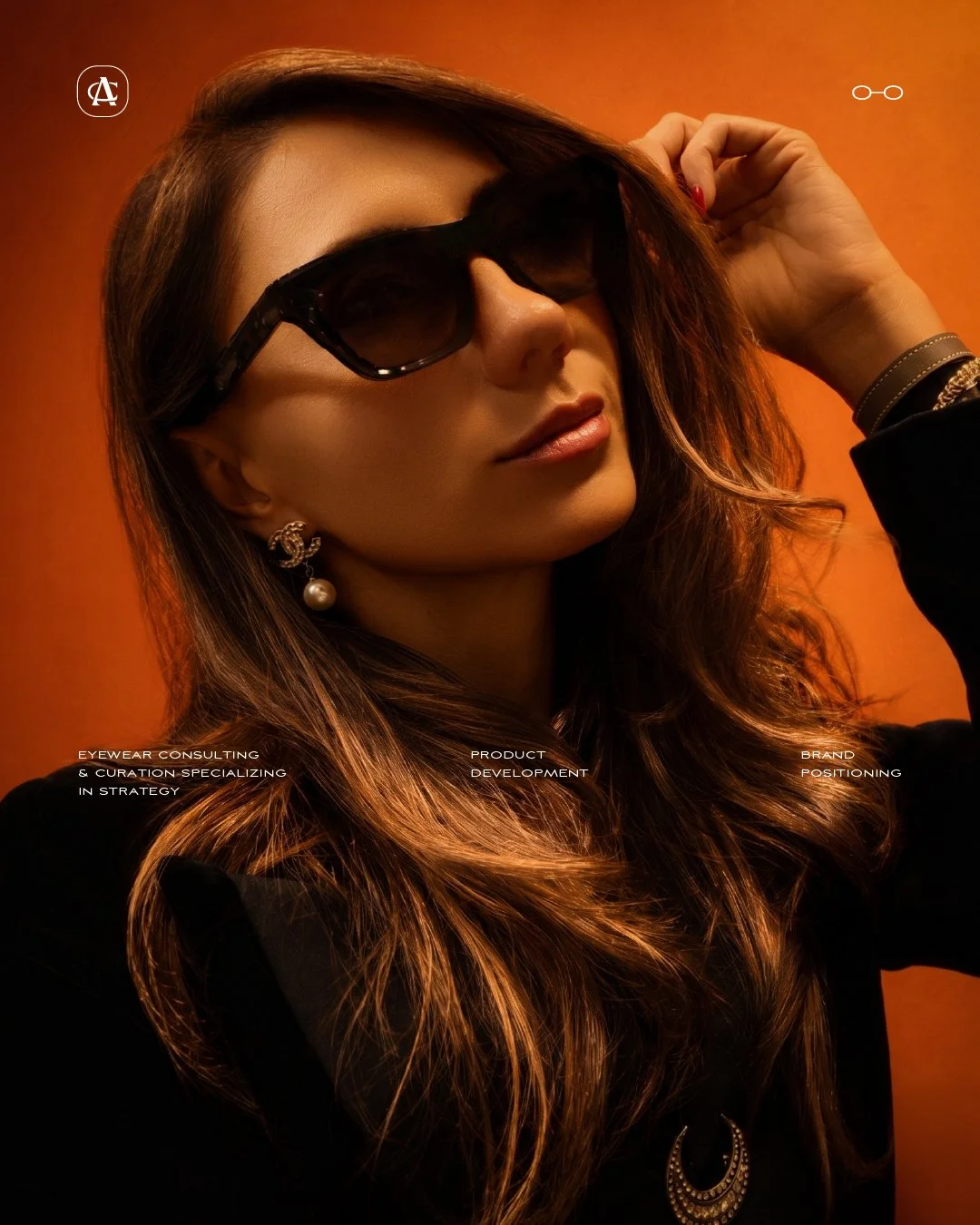 A woman with long, wavy brown hair wearing dark sunglasses, pearl earrings, a black outfit, and layered bracelets, posing against an orange background.