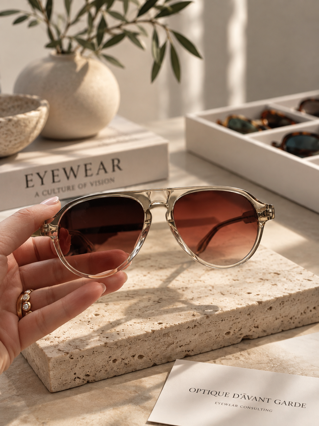 A hand holds a pair of sunglasses with dark lenses and a clear frame on a textured stone surface. Nearby are other sunglasses in a white tray, a book titled 'EYEWEAR: A CULTURE OF VISION,' a potted plant, and a piece of paper with French text about eyewear consulting.
