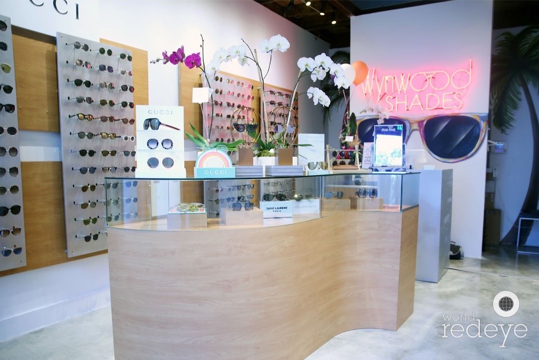 Display of sunglasses and eyeglasses at Wynwood Shades store with a neon sign, flowers, and a glass counter.