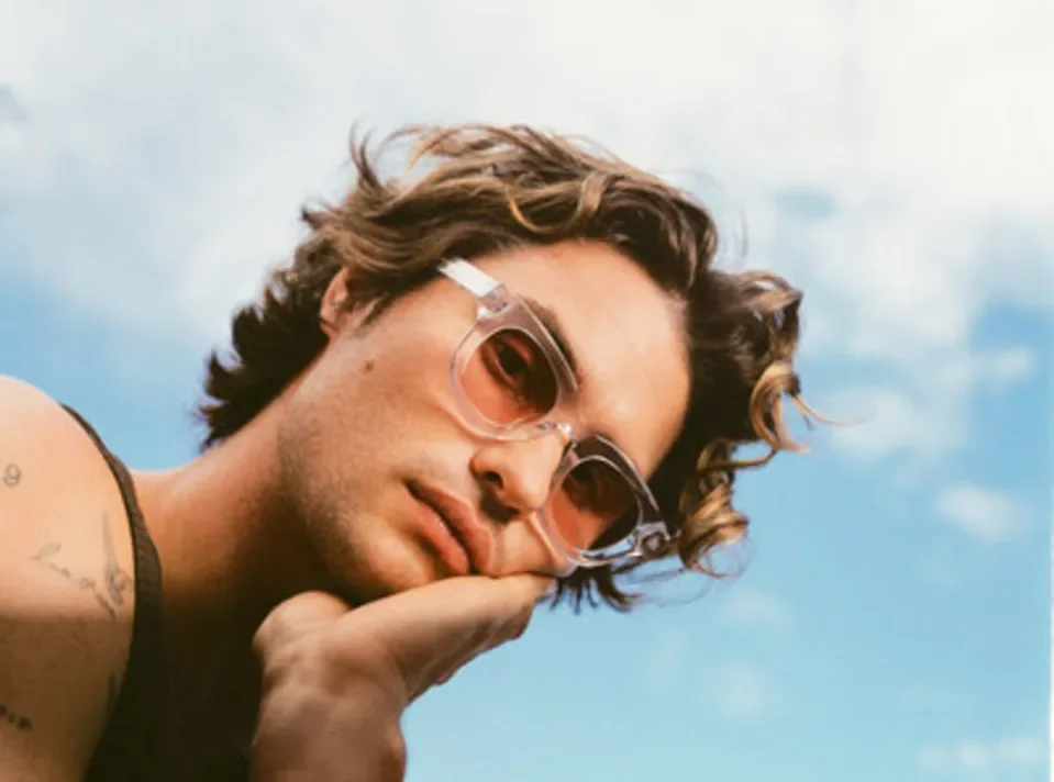 Close-up of a person with curly hair wearing sunglasses, resting their head on their hand against a cloudy sky background.