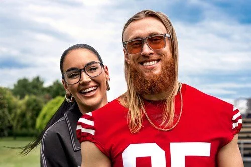 A smiling woman and man with long hair and beard, wearing sunglasses and casual clothing, standing outdoors on a cloudy day.