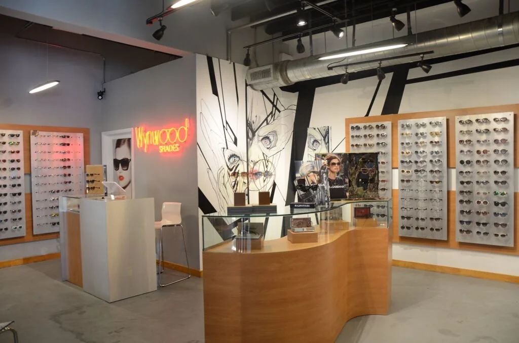 Interior of an eyewear store with display walls of sunglasses, a wooden counter, and a neon sign that says "Wynwood Shades".