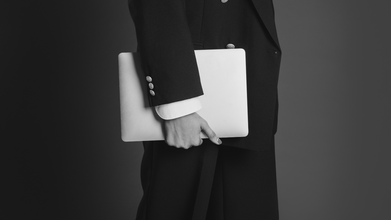 Marketing professional dressed in a suit holding a closed laptop against a plain background. No face is visible, walking off frame.