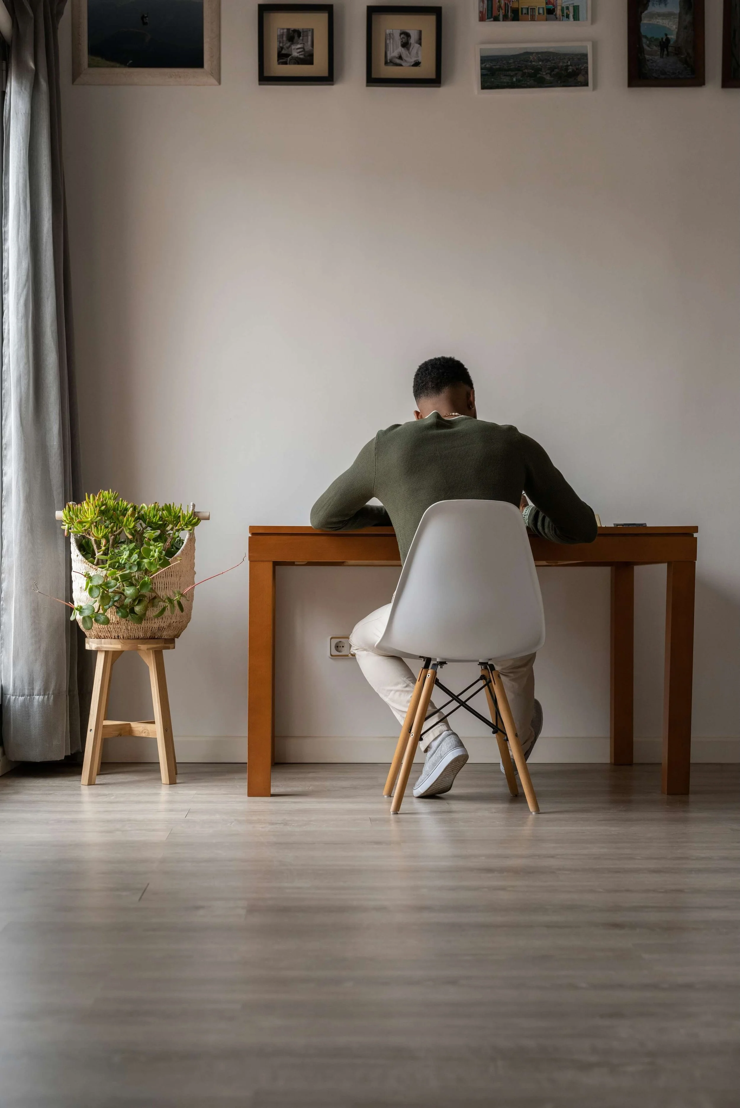 A person with short curly hair sitting at a wooden desk in a room with white walls, decorated with framed pictures and artwork, a gray curtain on the left, a potted plant on a wooden stand to the left of the desk, and a white chair with wooden legs.