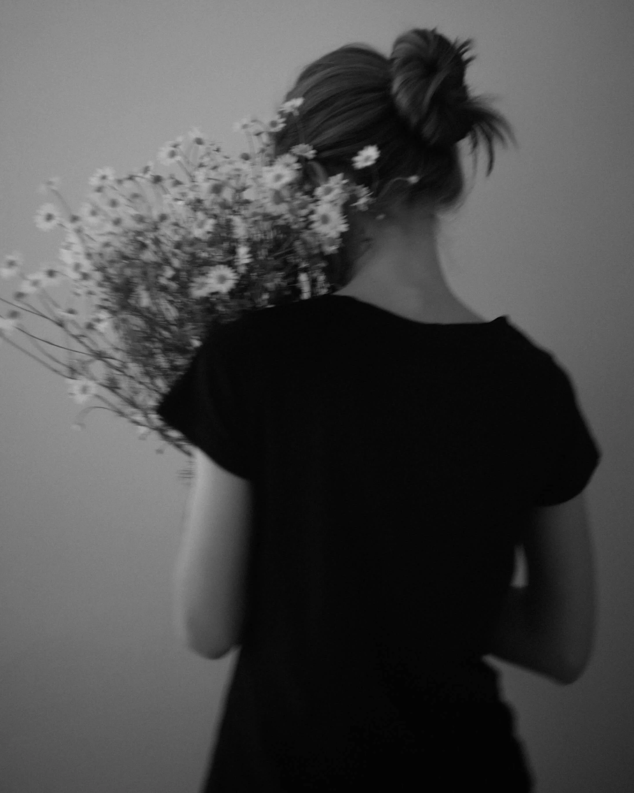 A person with dark hair styled in an updo, looking away, with a bouquet of small daisy flowers, and wearing a dark clothing, in a monochrome setting.
