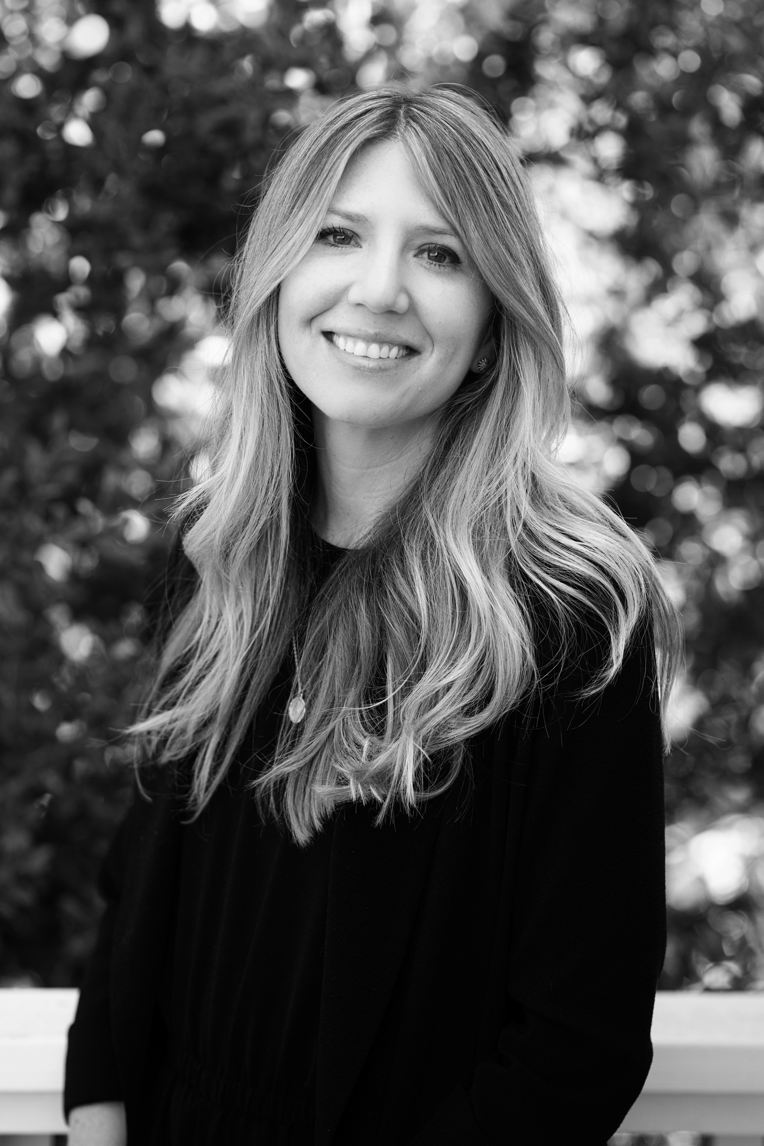 Portrait of a smiling woman with long, wavy hair, outdoors with blurred foliage in the background, in black and white.