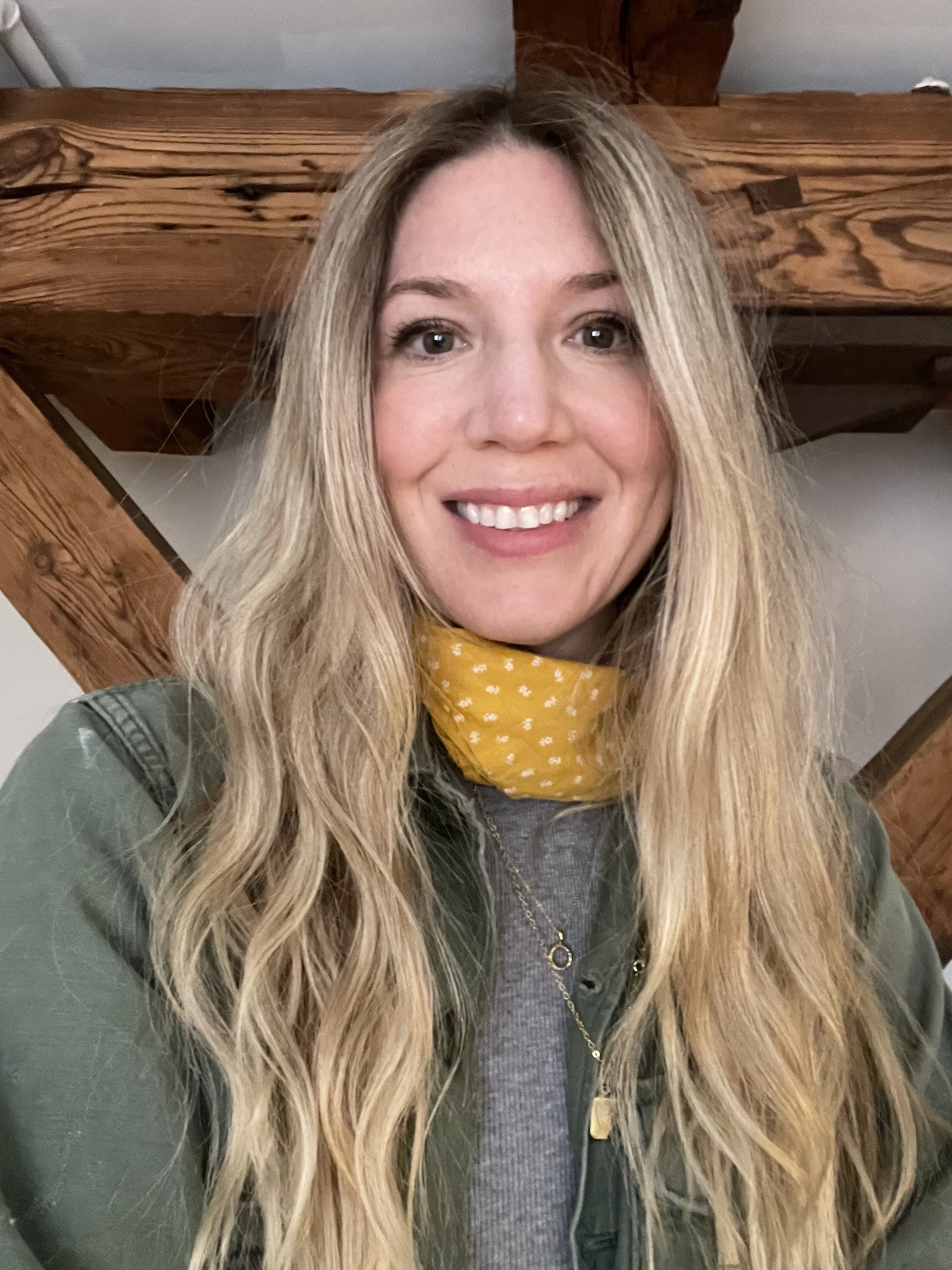 A woman with long blonde hair, smiling, wearing a yellow scarf with white polka dots, a gray shirt, and a green jacket, in front of a wooden staircase.