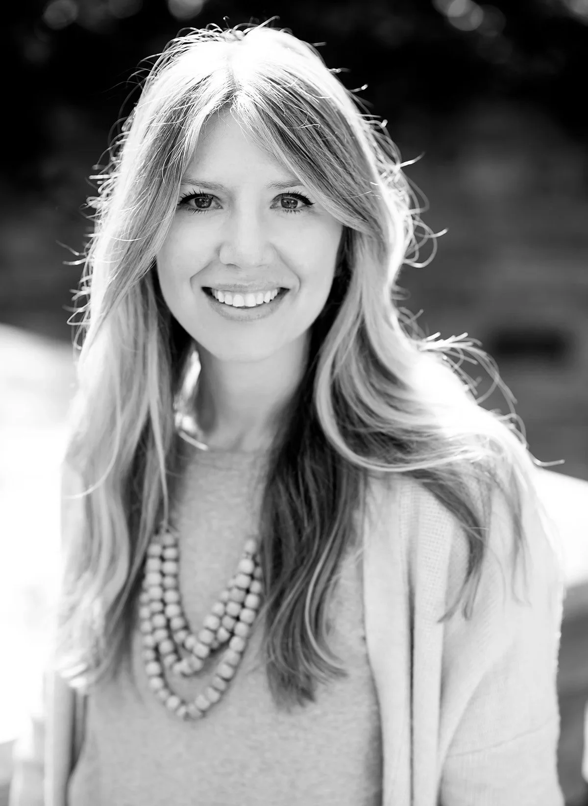 Black-and-white portrait of a smiling woman with long, wavy hair, wearing a beaded necklace, outdoors.