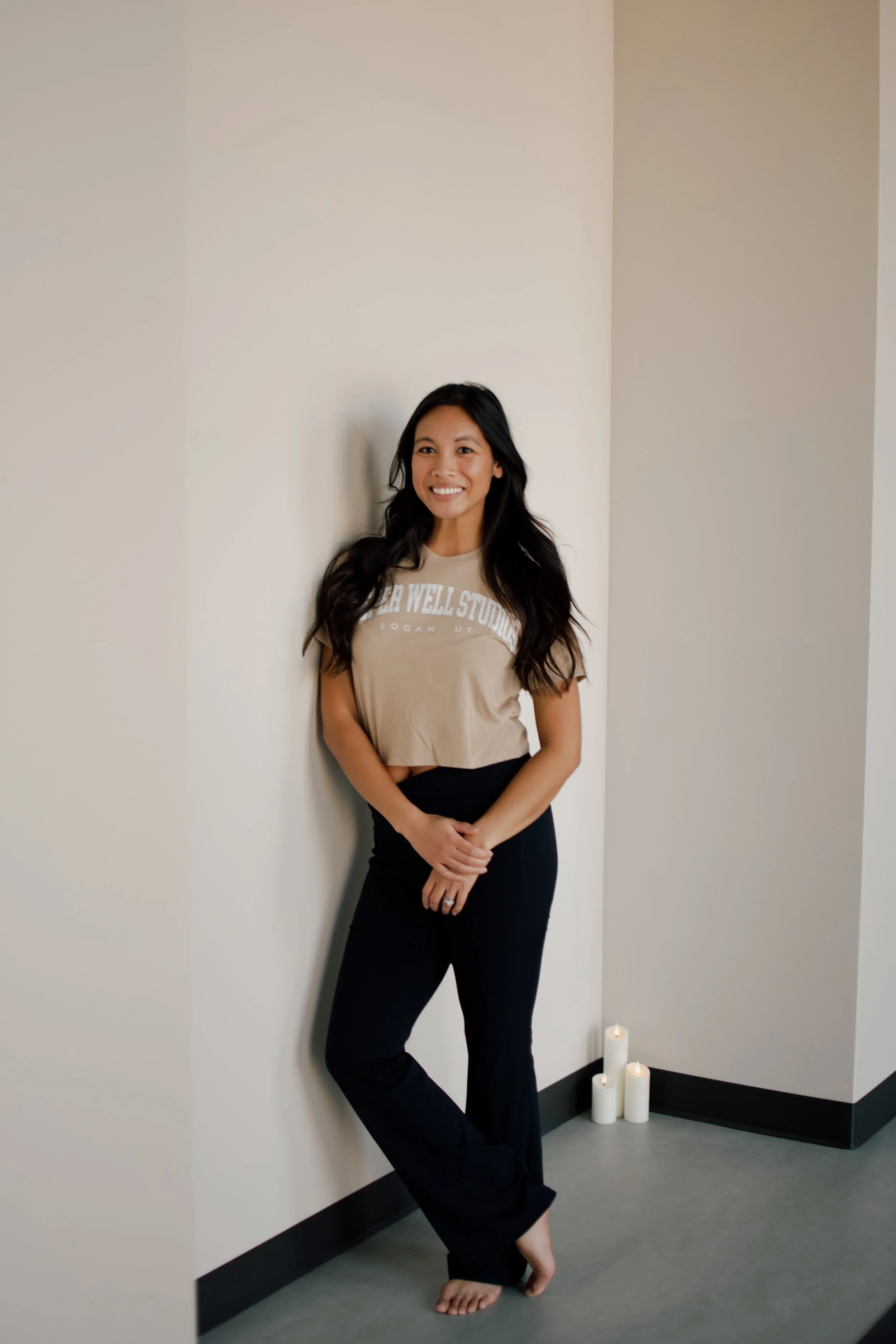 A woman with long, dark, wavy hair standing in a corner of a room with light-colored walls. She is smiling and wearing a beige t-shirt with white text and black pants. Candles are on the floor at the base of the wall behind her, and she is barefoot.
