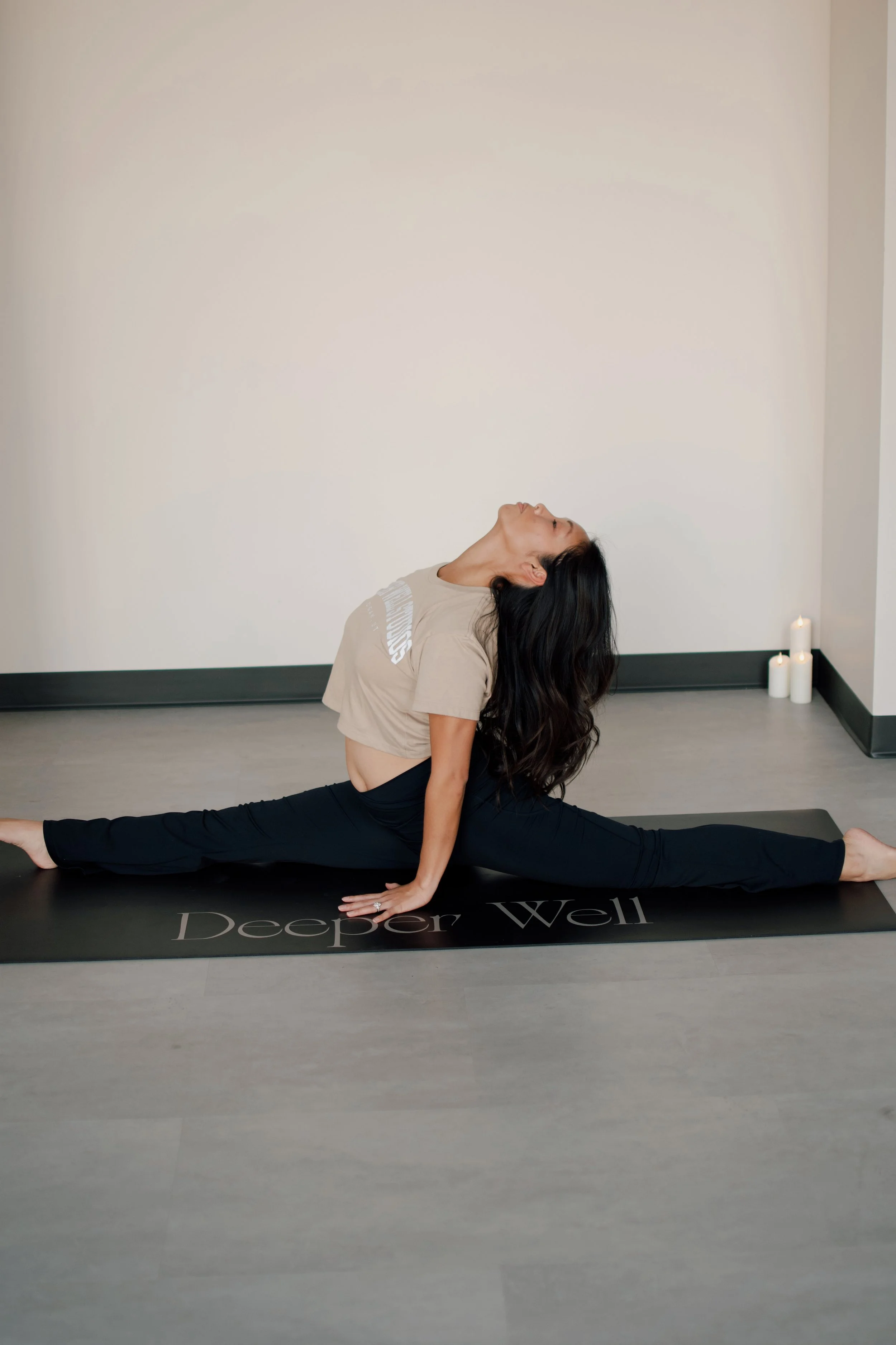 A woman practicing yoga in a split pose on a yoga mat in a minimalistic room with lit candles in the corner.
