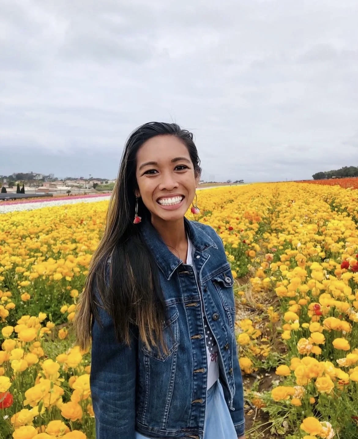 A young woman smiling in a field of yellow and orange flowers under a cloudy sky.