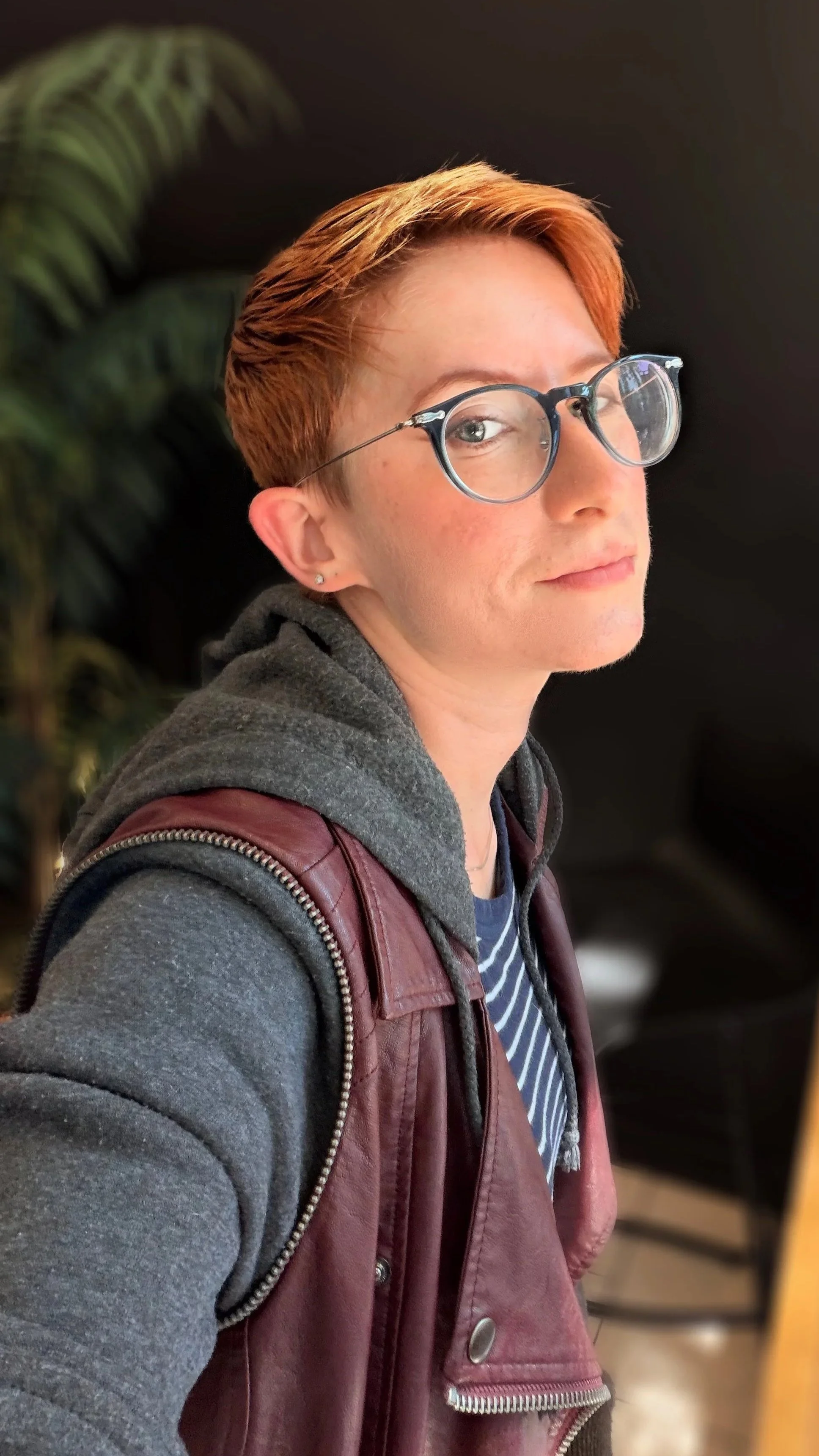 A young person with short reddish hair and glasses, wearing a grey hoodie, striped shirt, and a maroon leather jacket with a backpack, standing indoors near plants.