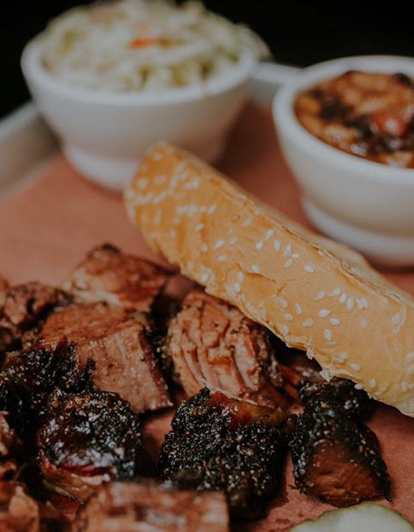 Barbecue platter with sliced smoked pork, barbecue sauce, a sesame seed bun, and side dishes in two white bowls, one filled with coleslaw and the other with baked beans.