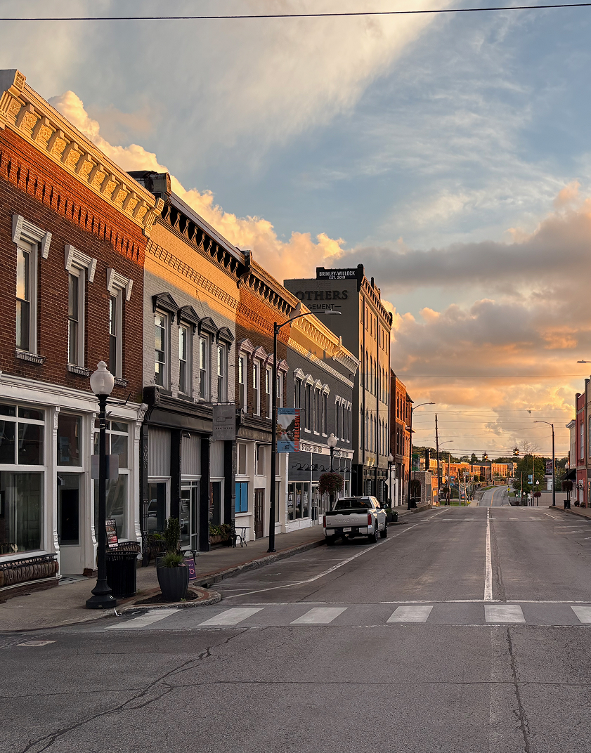 Empty street in a small town during sunset with buildings, parked cars, streetlamps, and a cloudy sky.