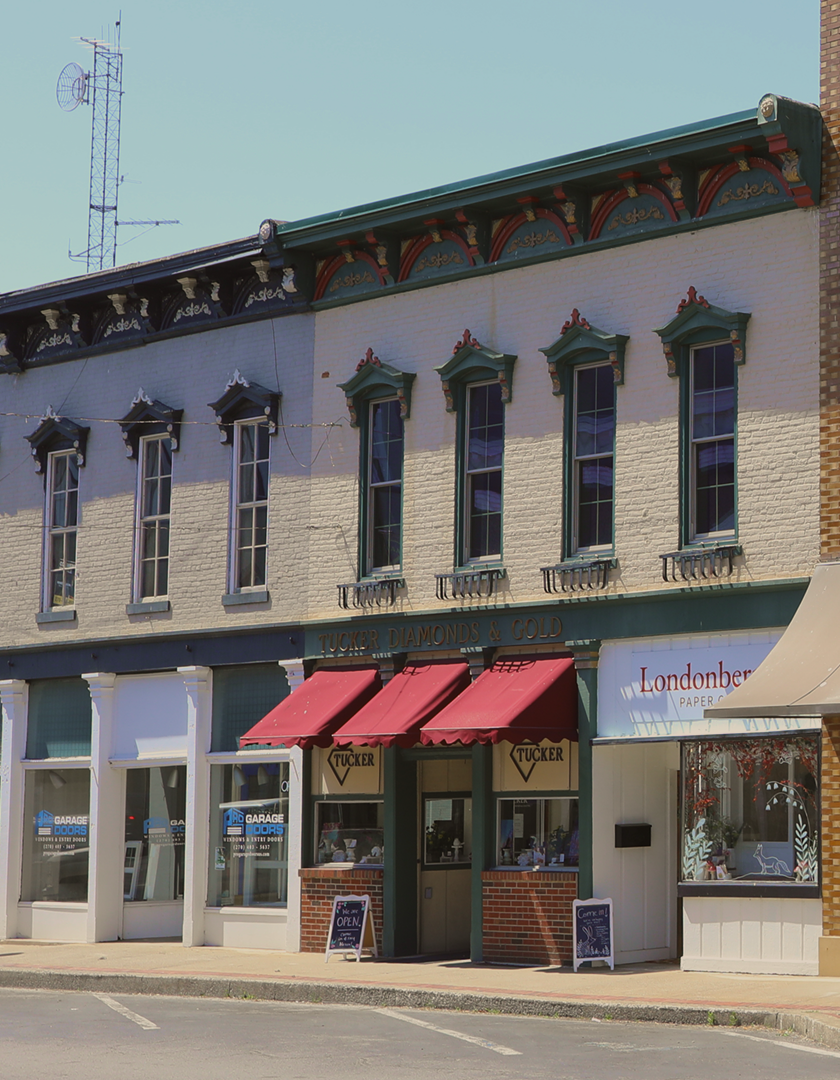 A two-story brick building with decorative green trim and red accents, featuring a storefront for Tucker Diamonds & Gold with red awnings. Adjacent to it is a store with signage for Londonberry Paper Co. The building has several tall, narrow windows with green frames. There is a parking lot in front.