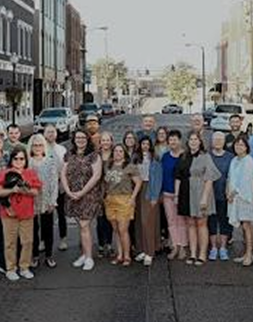 Group of people standing on city street with buildings, cars, and traffic lights in the background.
