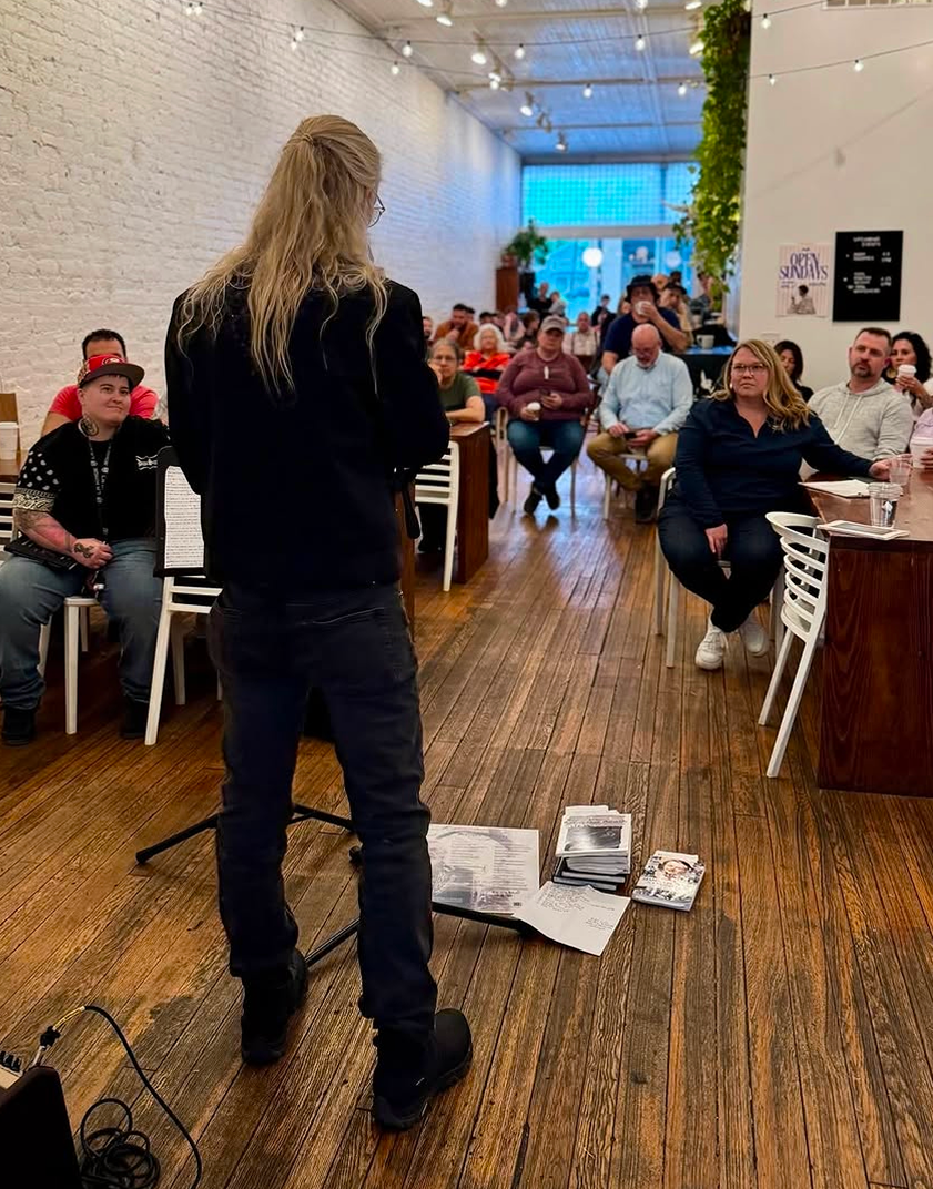 A woman standing in front of a seated audience in a coffee shop, giving a presentation or speech. The audience is attentively listening, some with drinks. The room has wooden floors, white brick walls, and string lights.