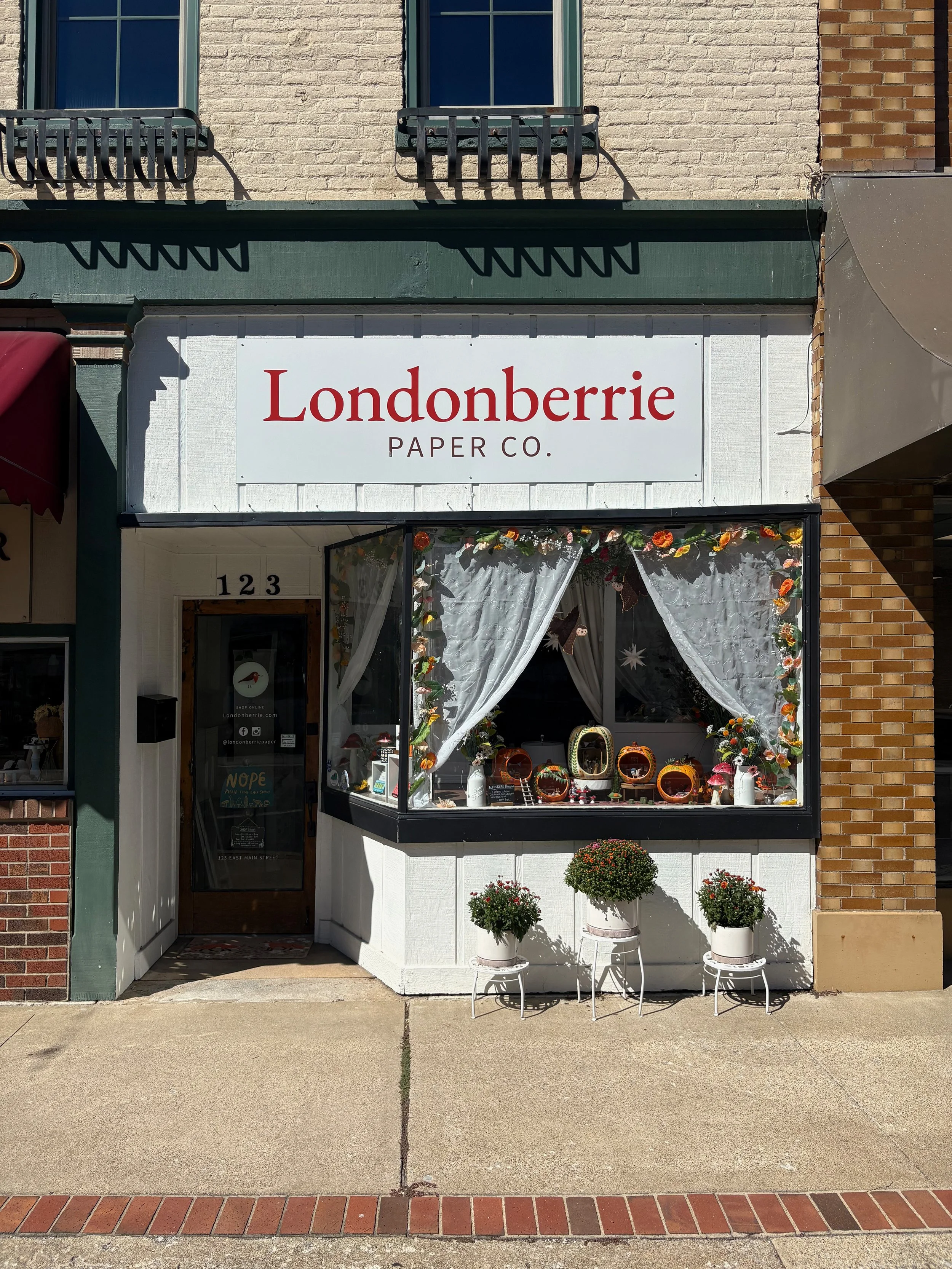Storefront with a sign that reads "Londonderrie Paper Co." with a display window showcasing decorated paper products, and two potted plants outside. The building has a brick and wooden exterior.