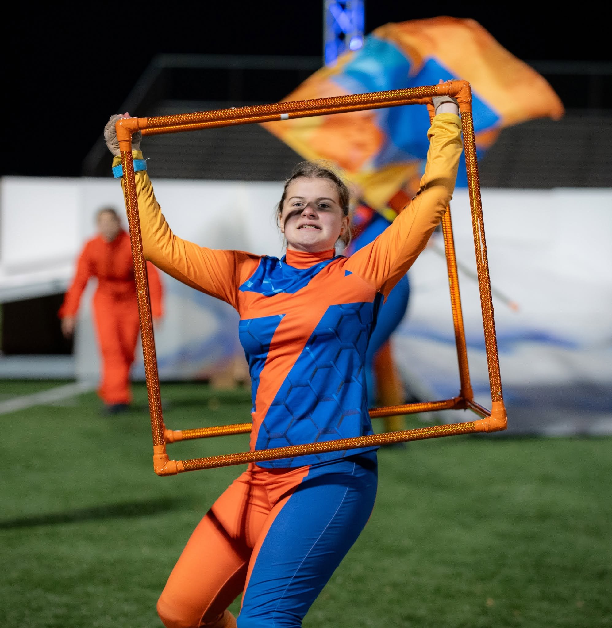 Female athlete in an orange and blue sports uniform holding a rectangular orange frame during a sporting event on a green field, with a blurred background of people and a large colorful flag.