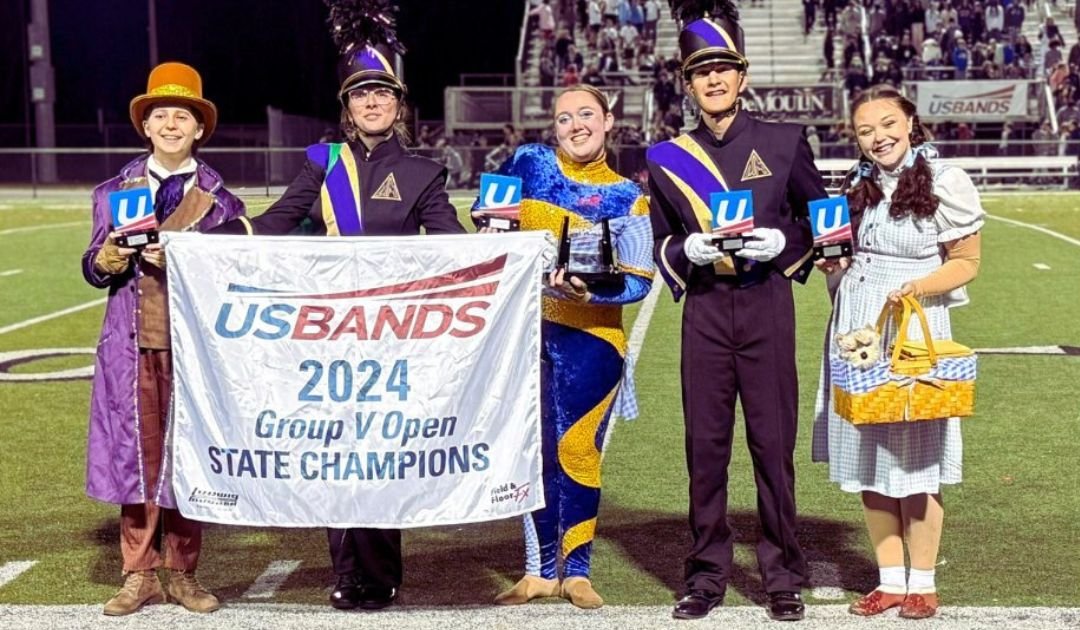 Group of five people in costumes standing on a football field holding a banner that reads 'US BANDS 2024 Group V Open State Champions.' They are dressed in colorful outfits, some with marching band uniforms, and are smiling while holding awards.