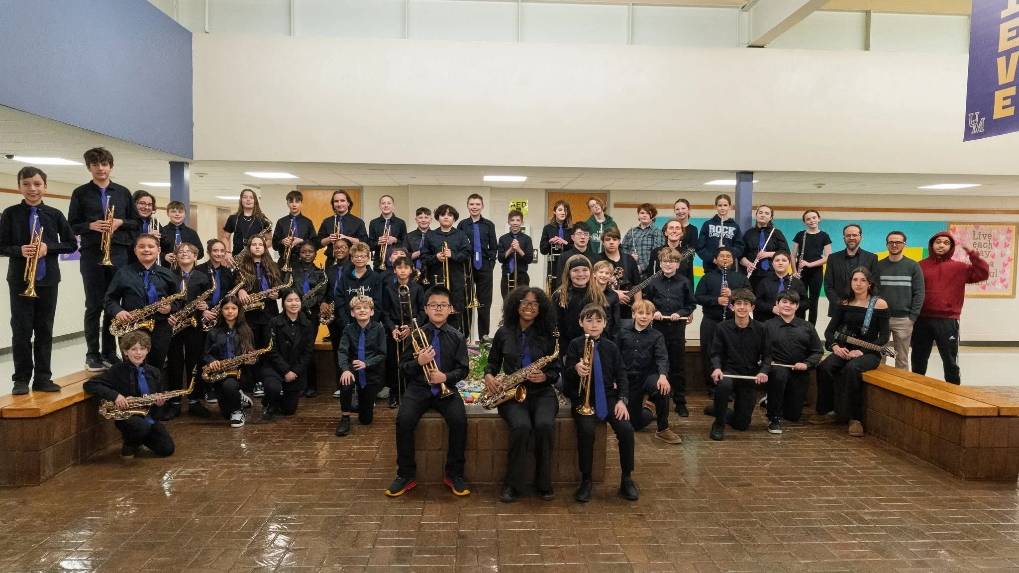 A large group of school students and teachers posing for a group photo in an indoor school setting, with some students holding musical instruments such as saxophones, trumpets, and guitars.
