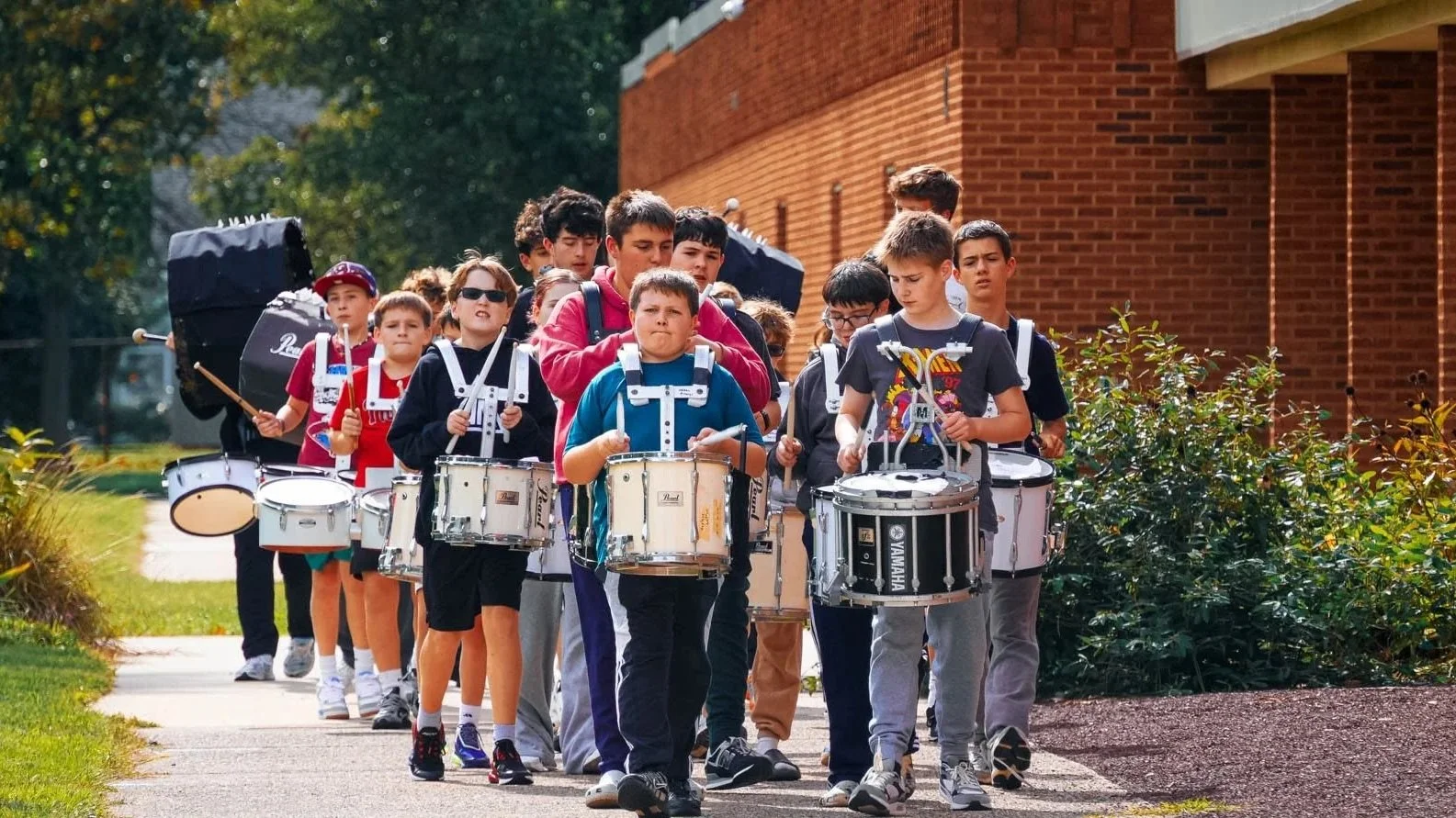 Group of young boys and teenagers marching on a sidewalk playing drums, led by a person with a large drum on their back, in front of a brick building, with greenery on the side.