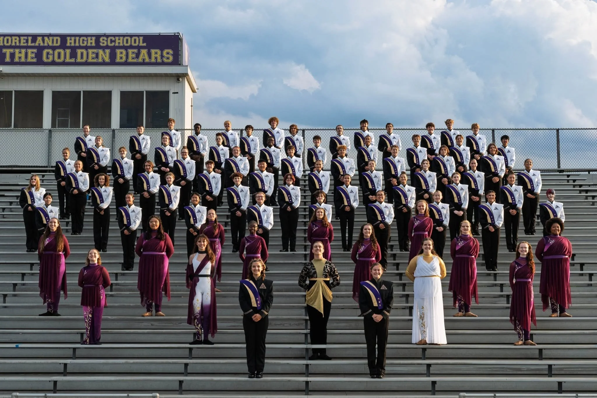 Group of students dressed in choir or band uniforms standing on stadium bleachers in front of a sign that reads 'Moreland High School The Golden Bears' with a cloudy sky in the background.