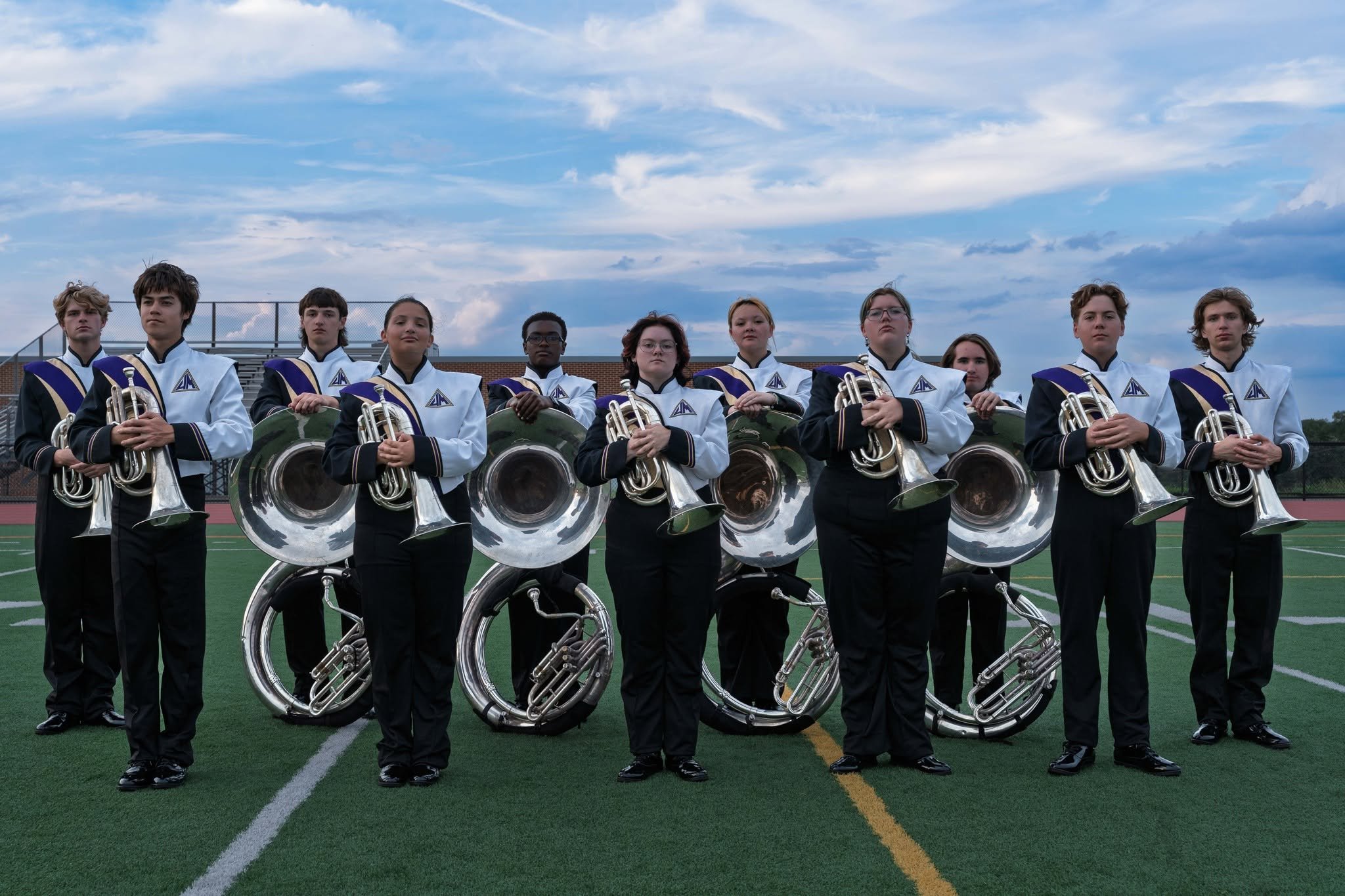 A marching band of young students dressed in white and black uniforms with purple and gold accents, holding brass instruments, lined up on a sports field under a partly cloudy sky.