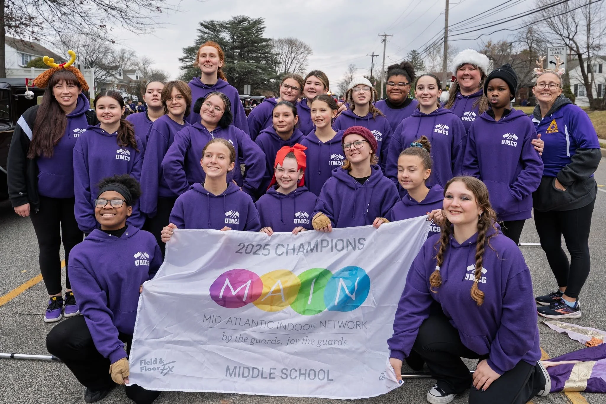 A group of young girls and women dressed in purple hoodies with UMGC logos, gathered outdoors during a celebration, holding a banner that reads '2025 Champions' and 'Middle School.' Some are wearing festive holiday accessories like reindeer antlers and Santa hats, and they are smiling and posing for the photo on a cloudy day.