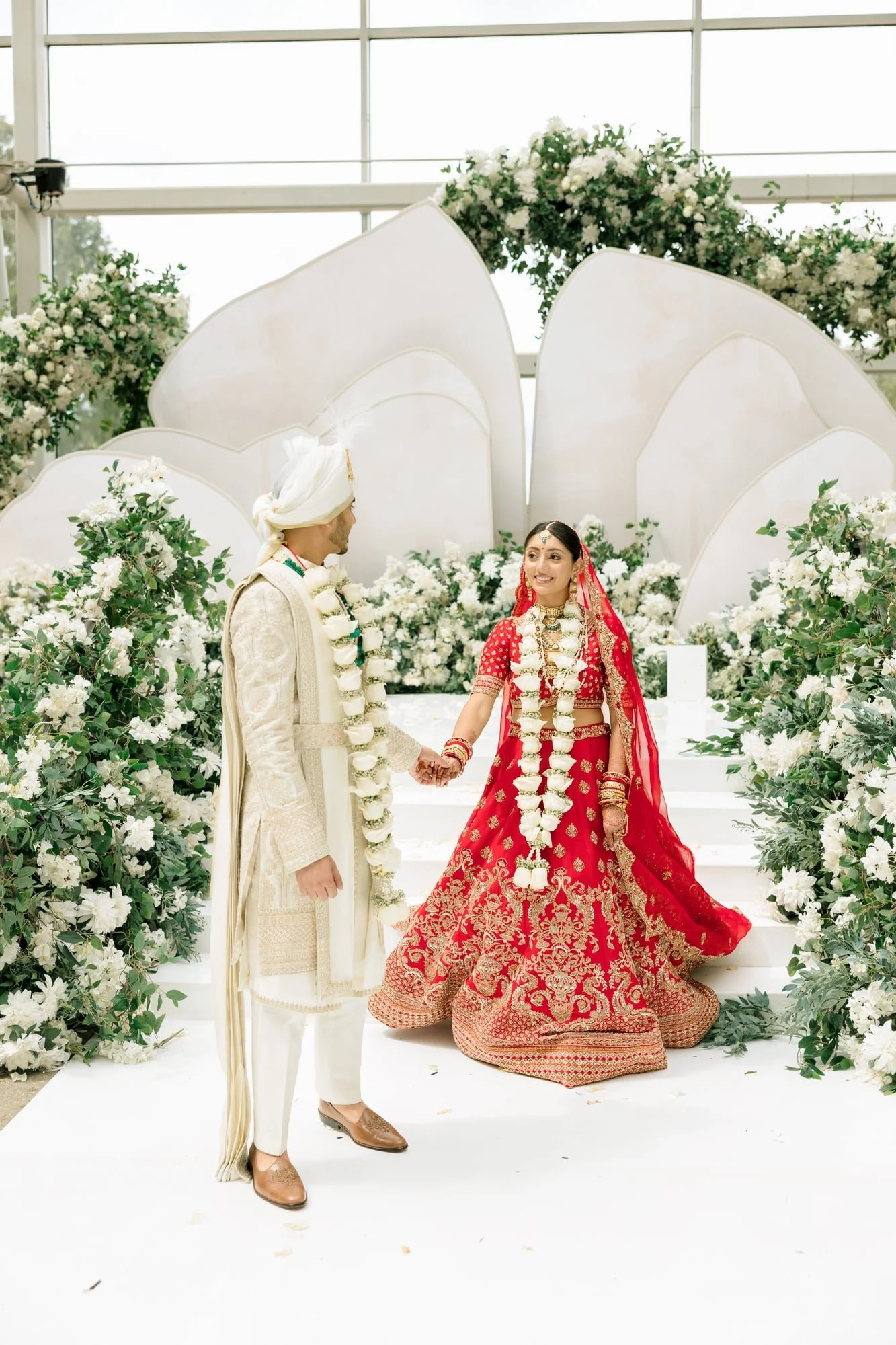 A bride and groom in traditional Indian wedding attire holding hands during their wedding ceremony, surrounded by white floral decorations.