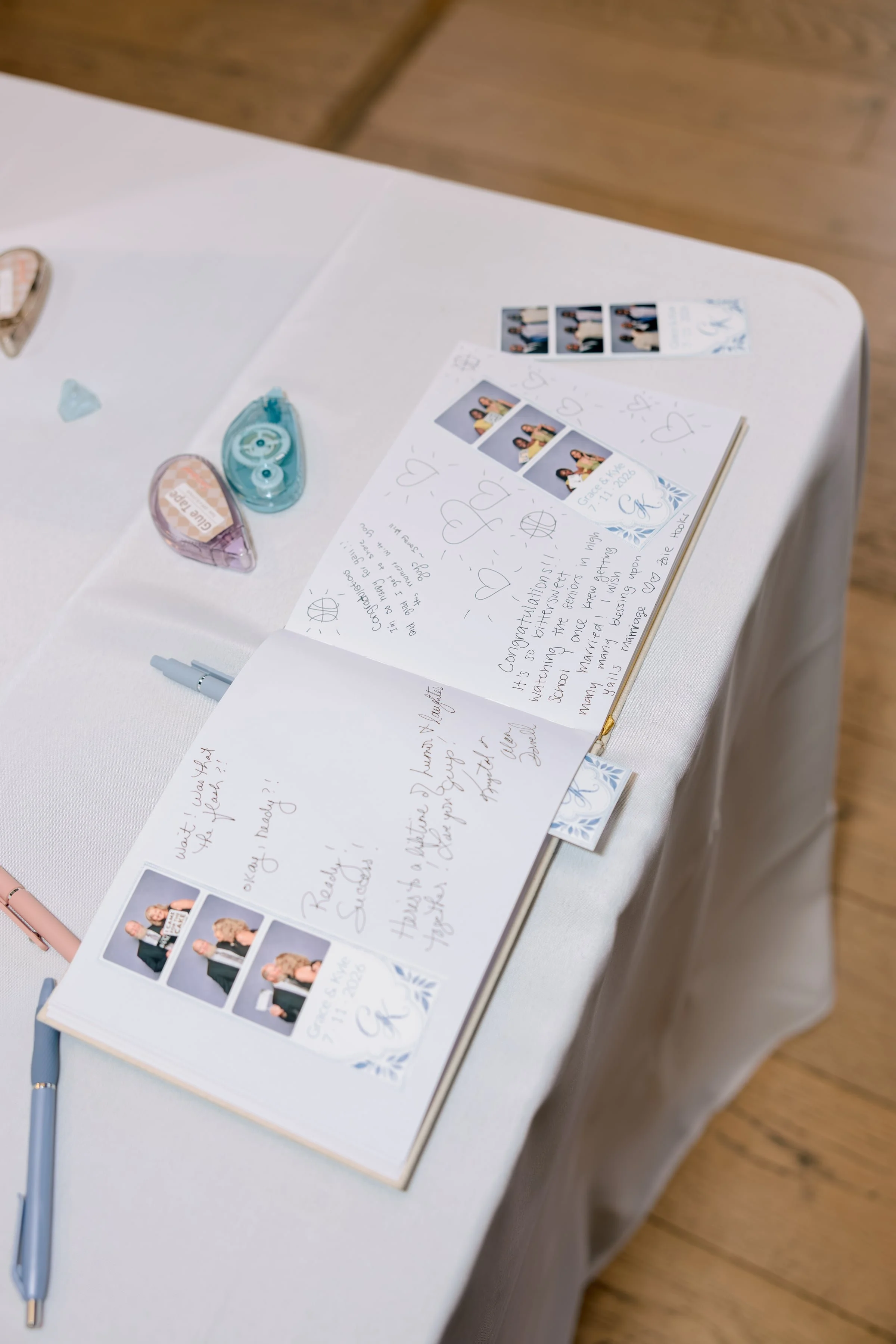 A wedding guestbook table with photo strips, handwritten notes, two photo guides with pictures of people, a gray pen, and small decorative objects including a blue transparent item and a pink item.