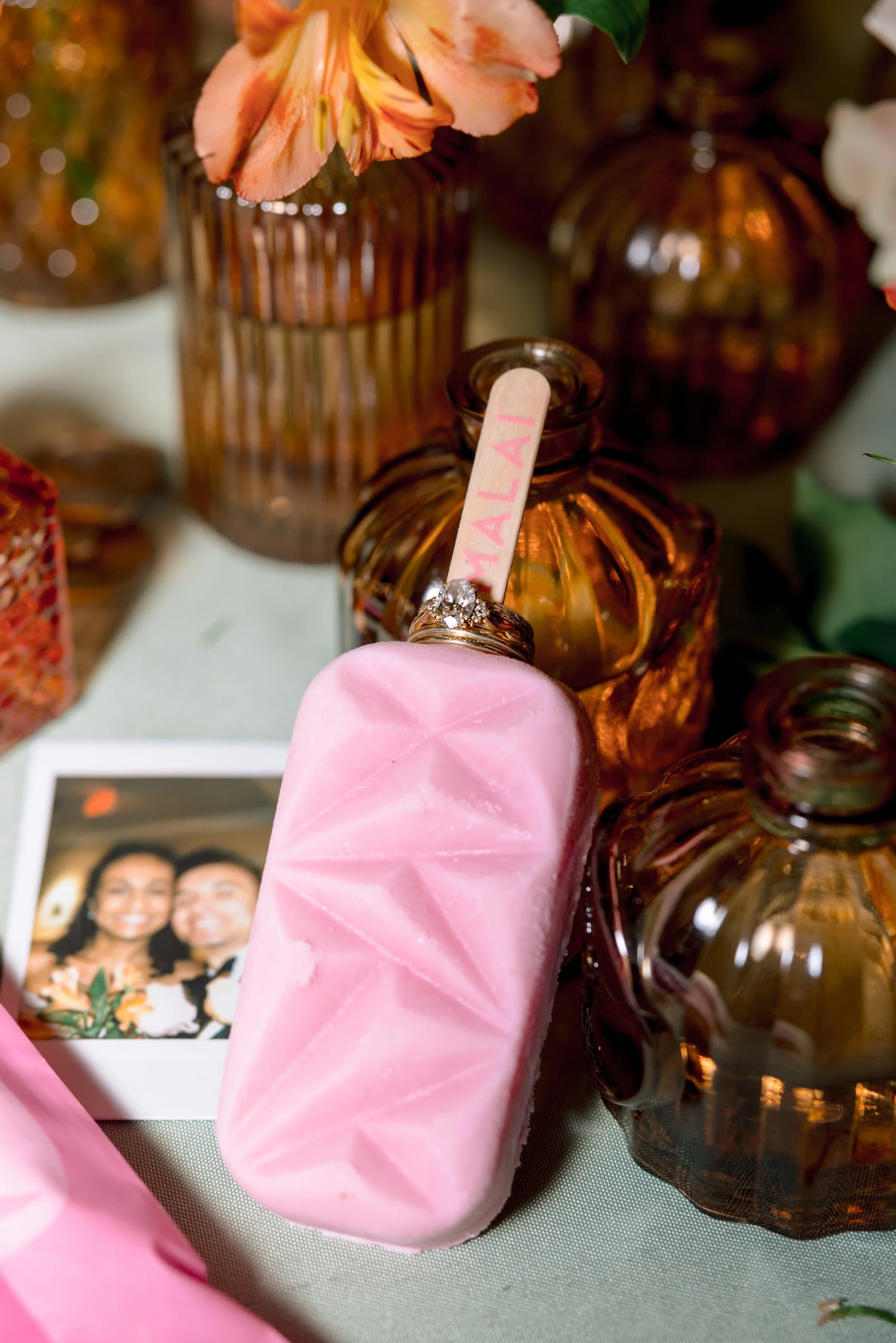 Close-up of pink soap with a ring, surrounded by amber-colored glass bottles and a framed photograph of a smiling couple.
