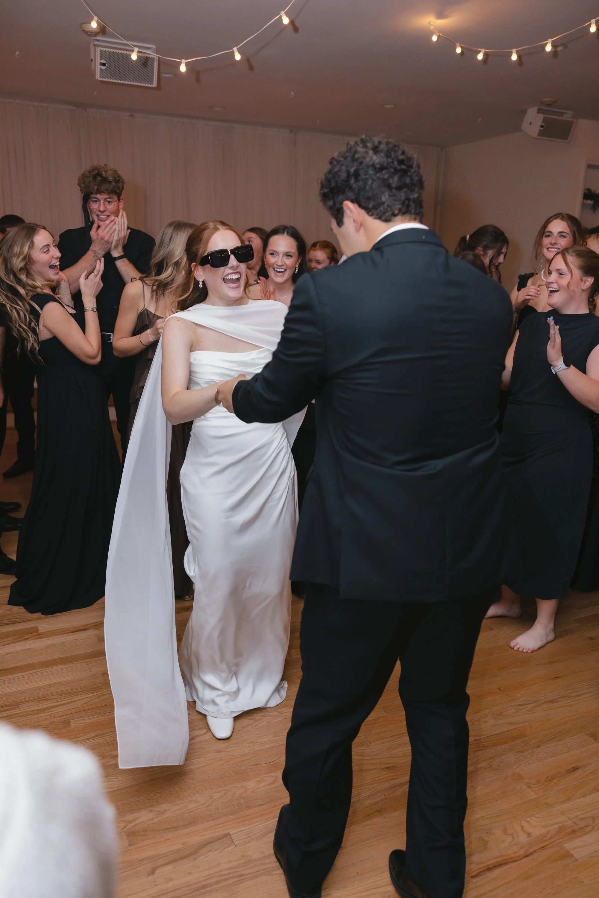 A group of people dancing and celebrating at a wedding reception, with the bride and groom in the center, smiling and holding hands. The guests are joyful and laughing, dressed in formal attire, some with glasses. The setting has wooden floors and st