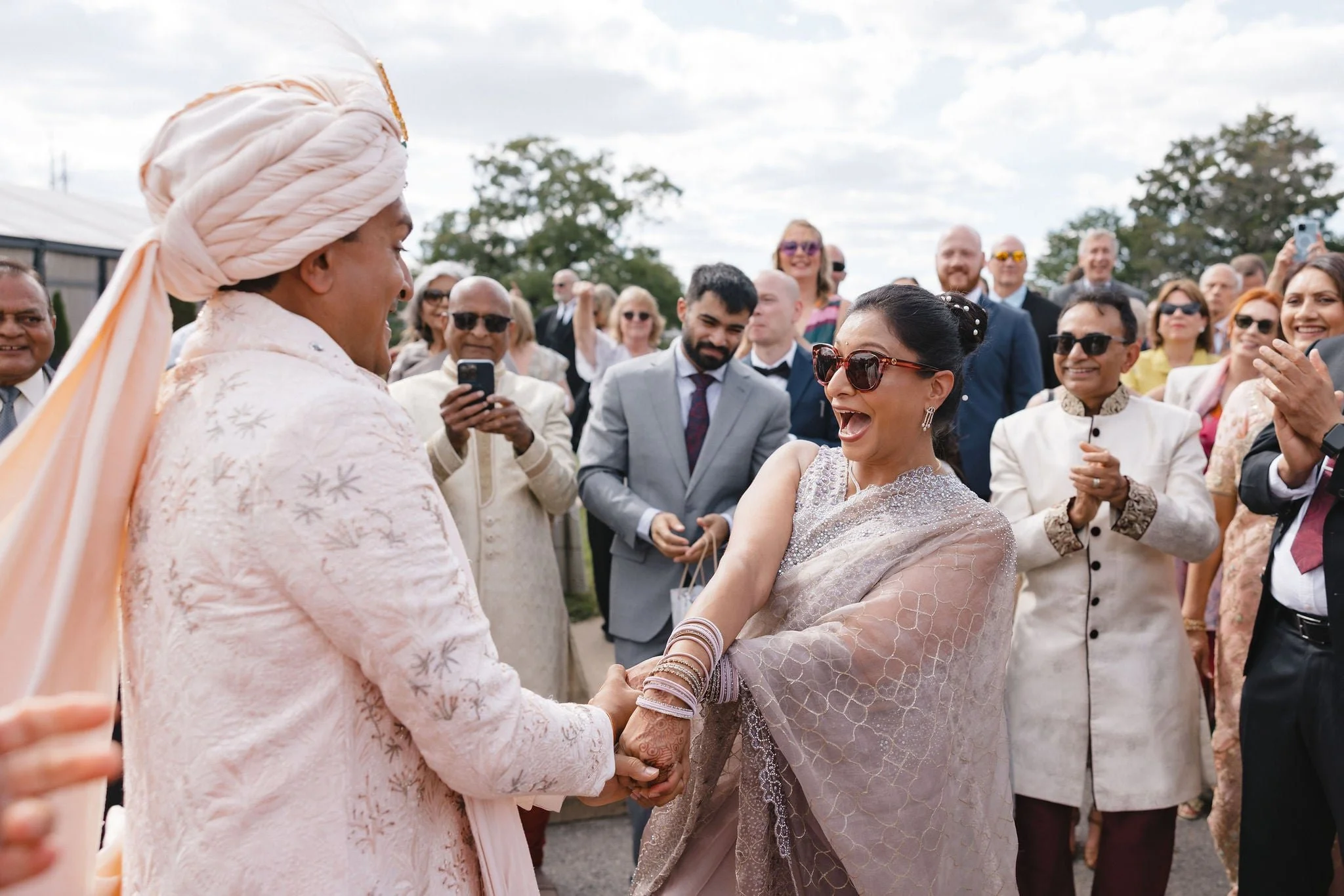 A woman and a man in traditional South Asian attire holding hands, surrounded by smiling and taking photos of a group of people outdoors during a celebration or wedding ceremony.