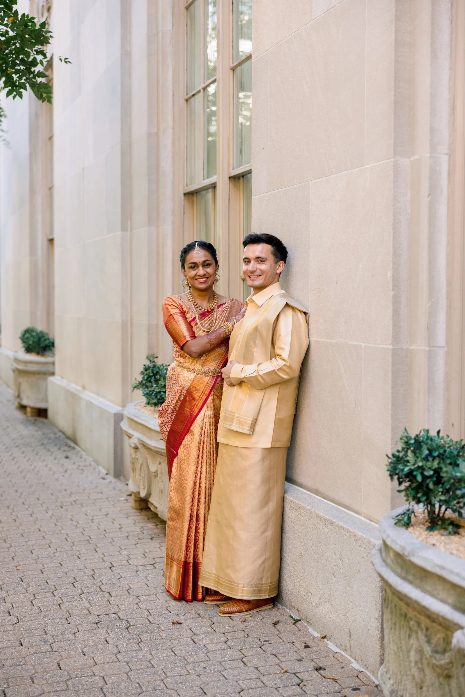 A South Asian couple dressed in traditional clothing leaning against a beige stone wall in an outdoor setting. The woman is wearing an orange and gold sari, and the man is in a cream-colored kurta with a matching dhoti. There are potted plants on the