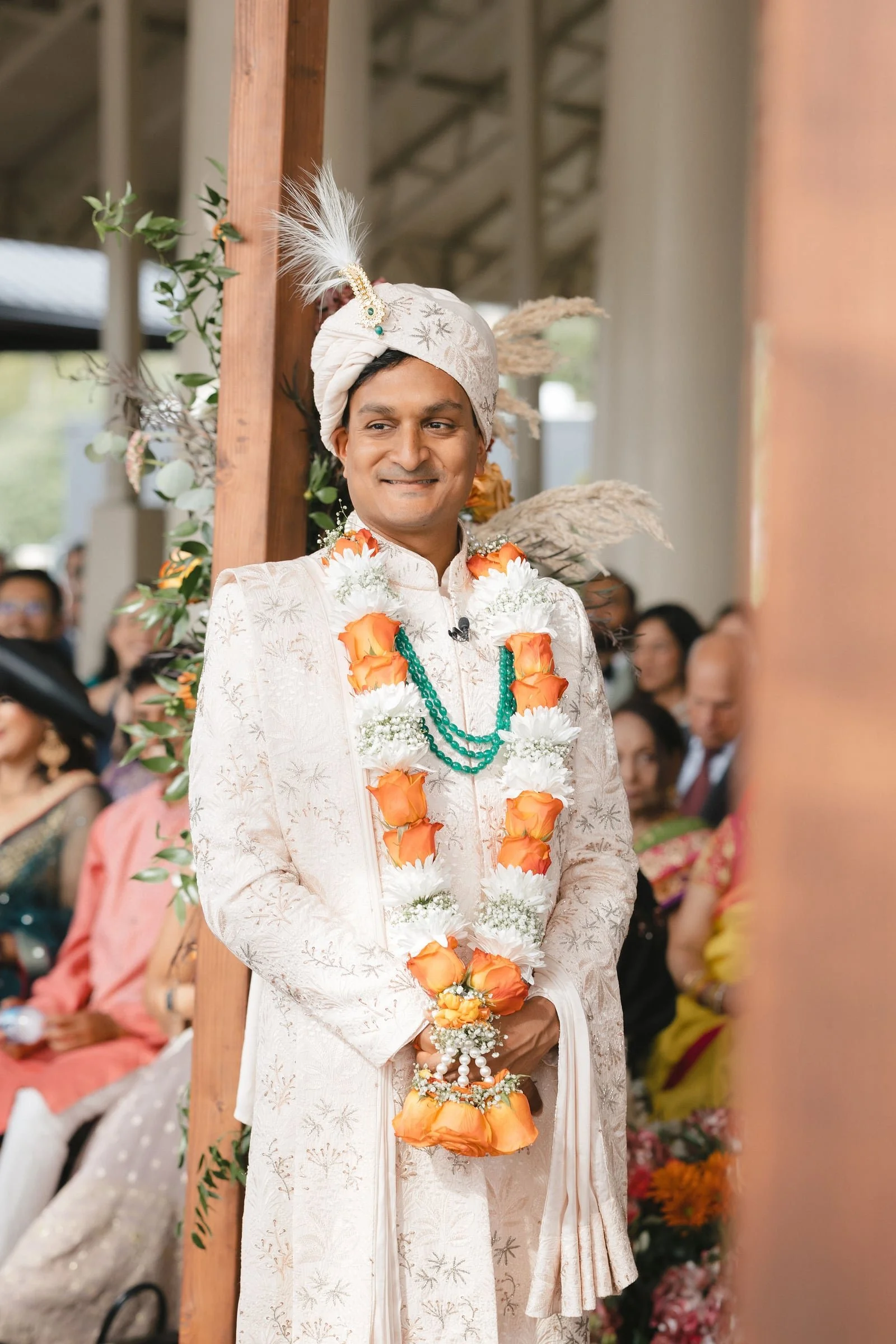 A groom dressed in traditional Indian wedding attire, wearing a cream-colored sherwani with intricate embroidery, a floral garland, and a turban with feathers. He is smiling and holding a floral accessory during a wedding ceremony, with guests seated